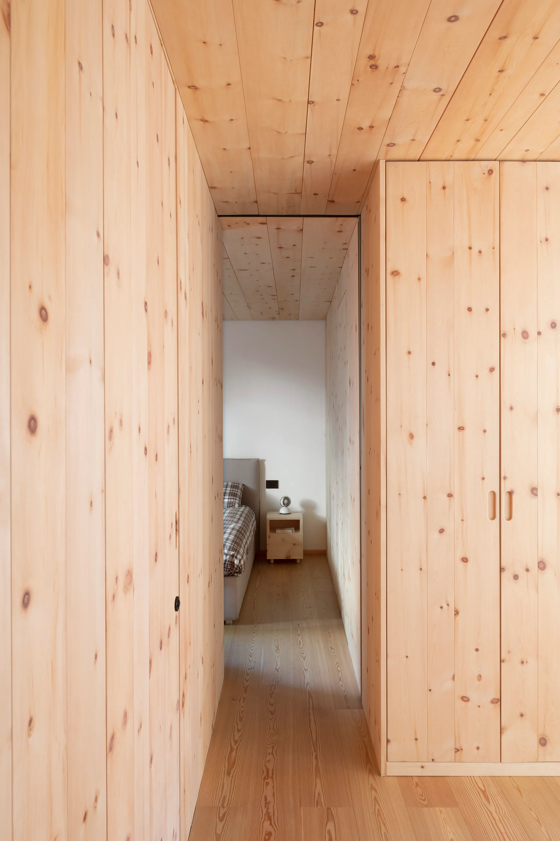 A hallway clad in knotty pine wood paneling on the walls and ceiling leads to a bedroom featuring a gray bed and matching wood nightstand.