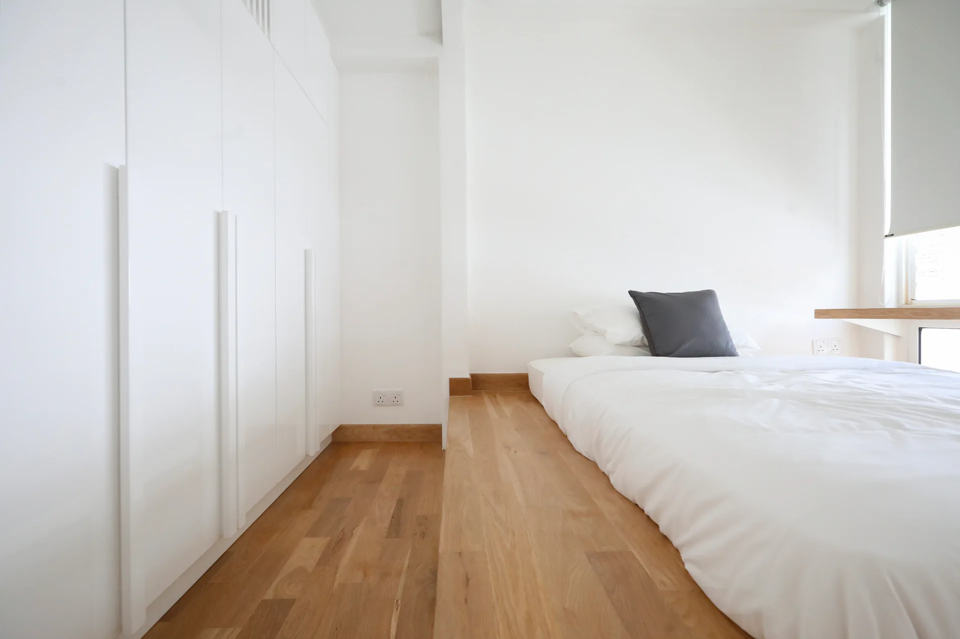 A minimalist white bedroom features a mattress on a raised light wood platform next to a tall, built-in white wardrobe.