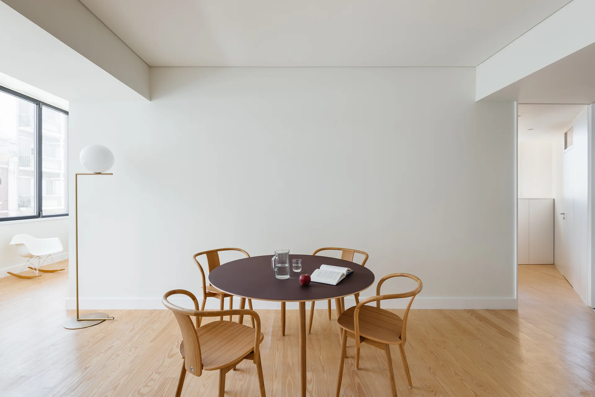 A minimalist dining area features a dark round table surrounded by four wooden chairs on light wood floors against a plain white wall.