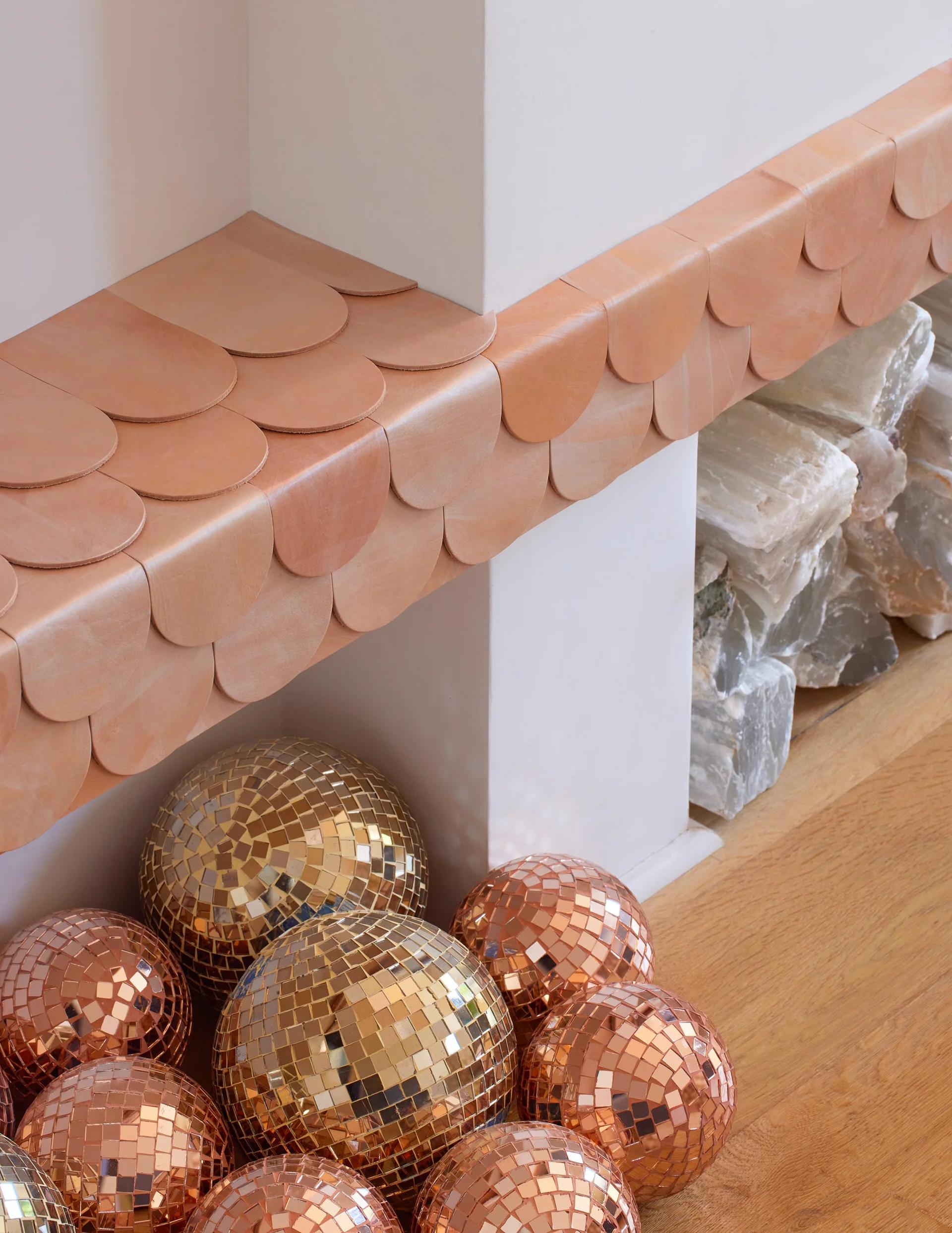 A close-up of a fireplace showing a mantel covered in scalloped tan leather tiles, with copper disco balls and white raw stones on the floor.
