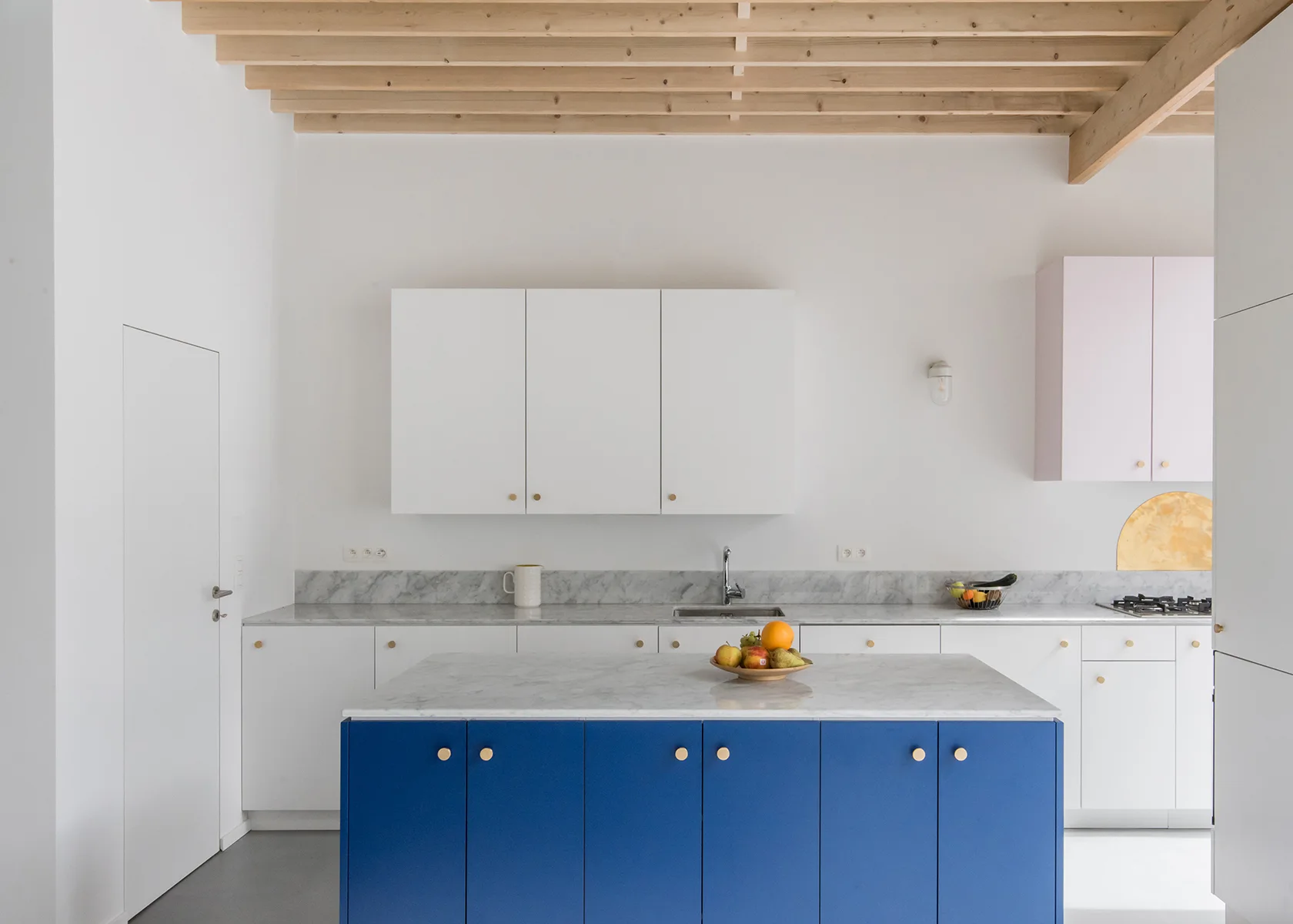 A minimalist white kitchen features a vibrant blue island with marble countertops, white cabinetry, and an exposed natural wood beam ceiling.