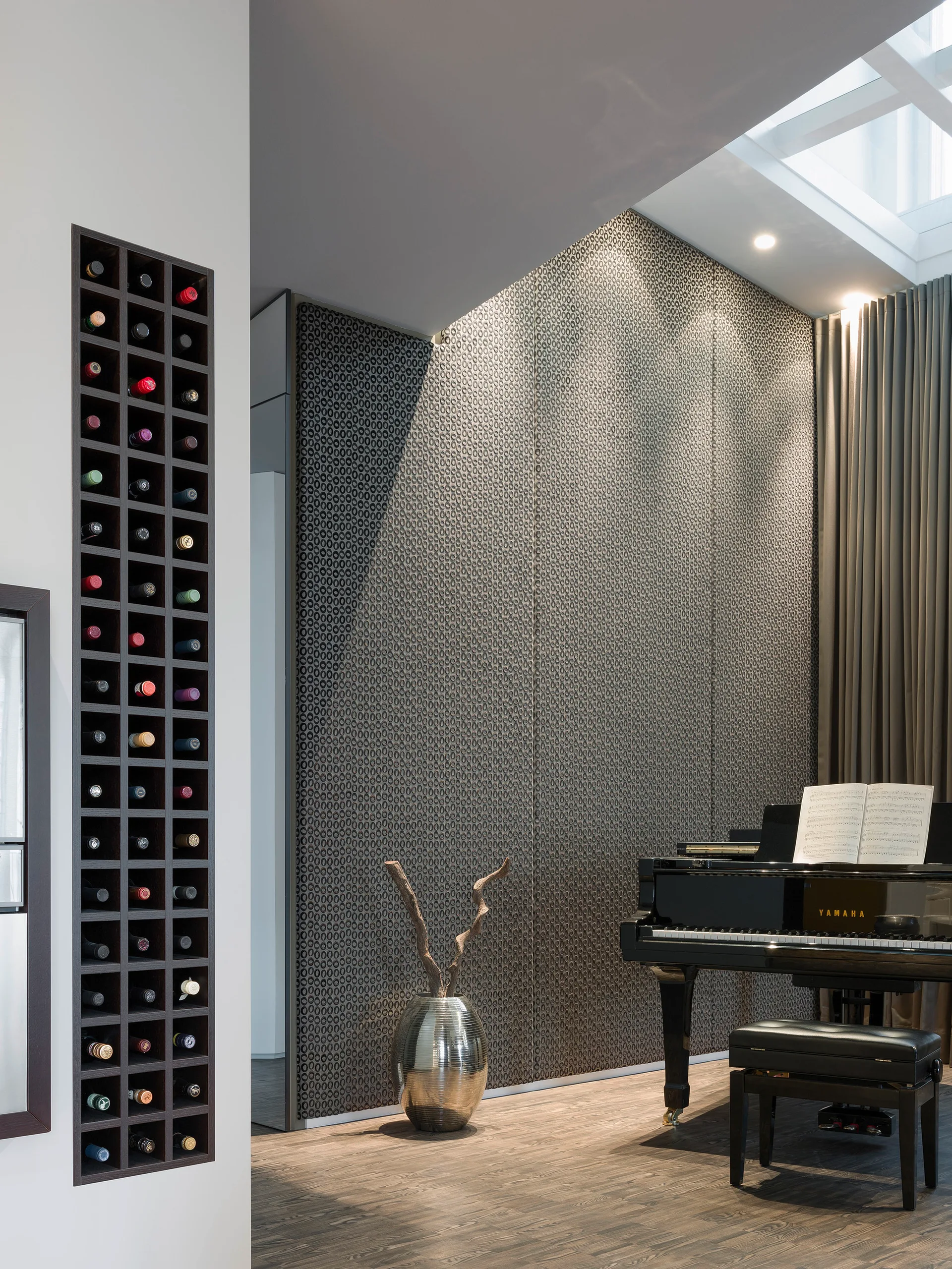 A black grand piano stands in a room with a grey patterned wall, a metallic floor vase, and a built-in dark wood wine rack.