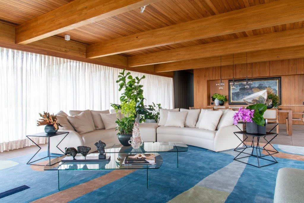 A contemporary living room features a cream sectional sofa and a blue patterned rug under a warm wood paneled ceiling with exposed beams.