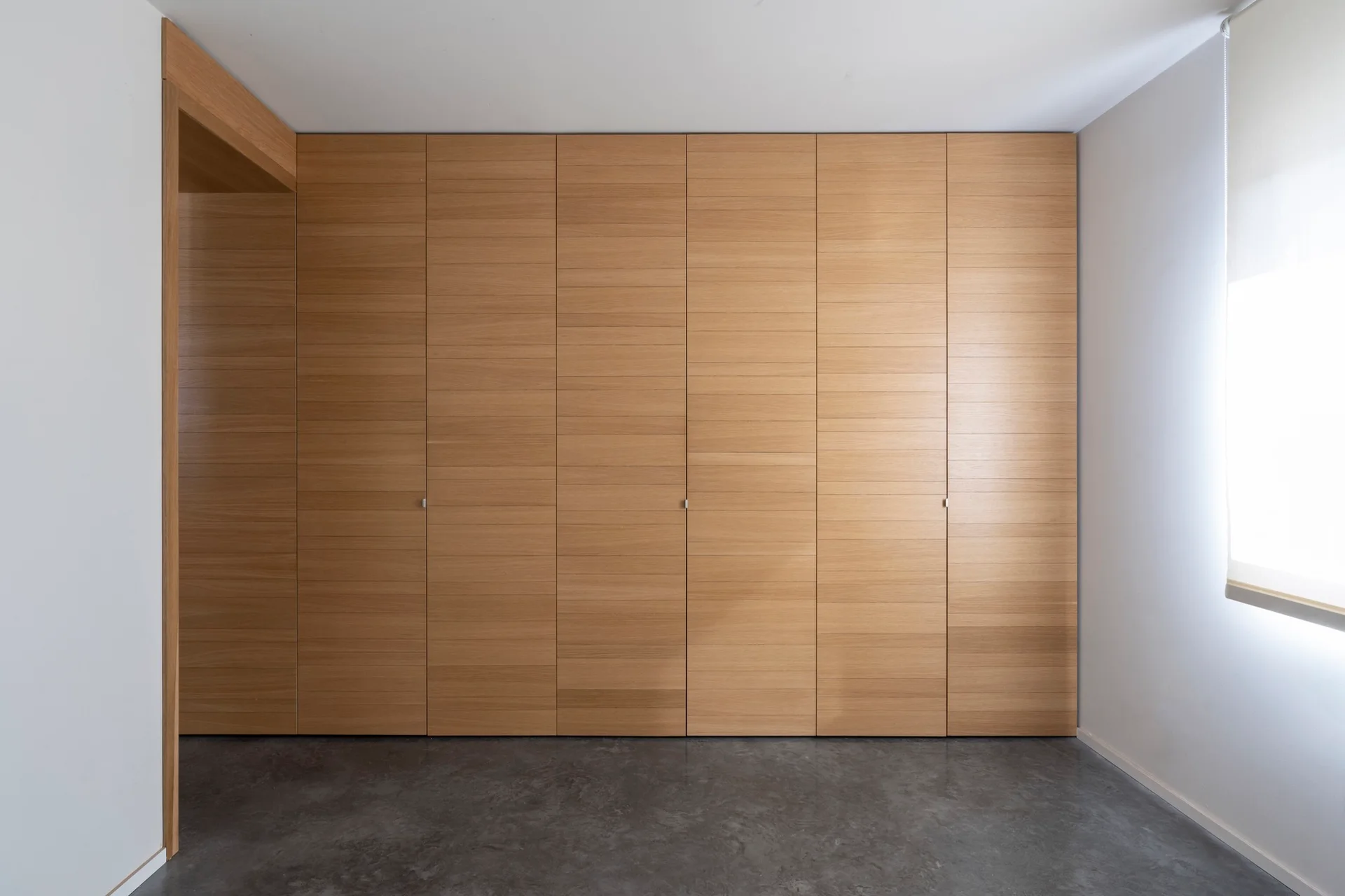Floor to ceiling light wood closet doors with horizontal grain span a wall in a room with a concrete floor and a window.