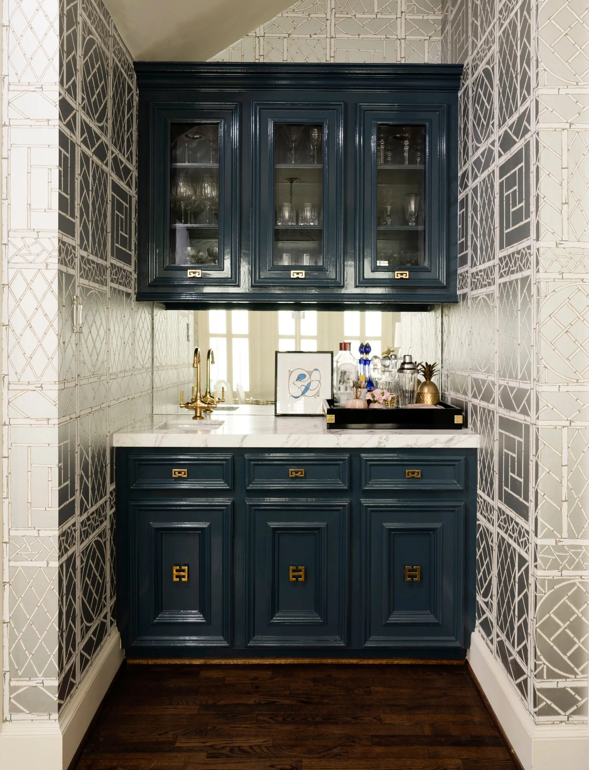 A compact home wet bar featuring high-gloss blue cabinetry with brass hardware, a marble countertop, and geometric patterned wallpaper.