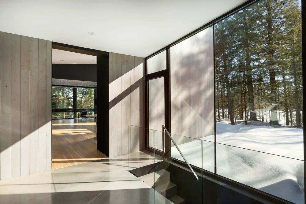 Minimalist entryway with vertical wood paneling, a glass staircase railing, and large windows looking out at a snowy forest.