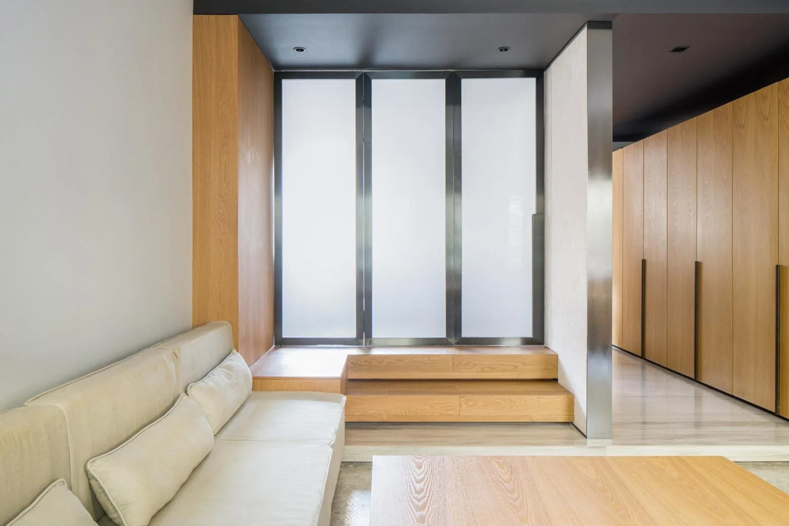 A minimalist living room featuring a beige sofa, a low wood table, and tiered wood platform steps leading to a large frosted window.