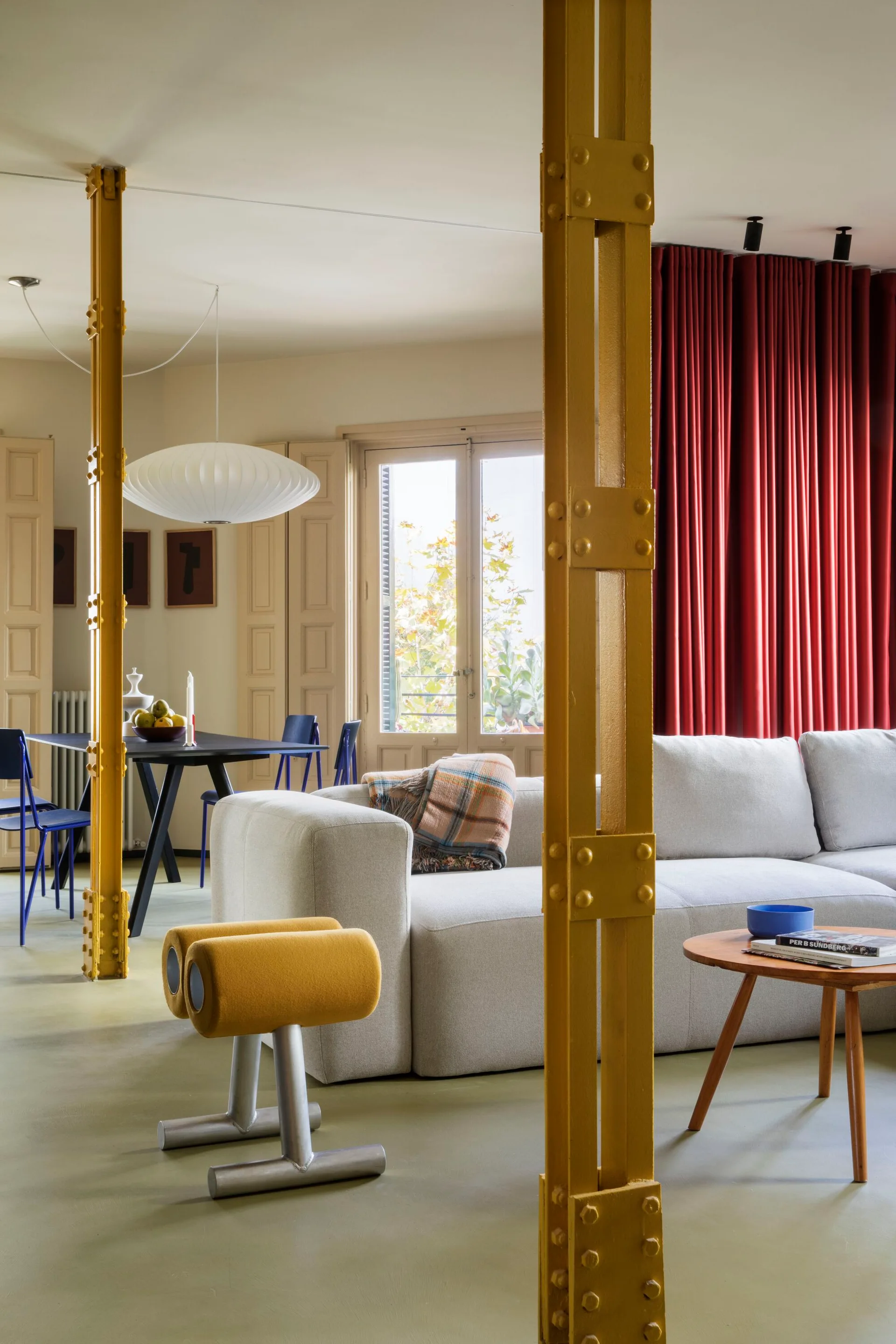 An open living and dining area featuring bright yellow steel columns, a neutral sofa, a bold red curtain, and a black dining table.
