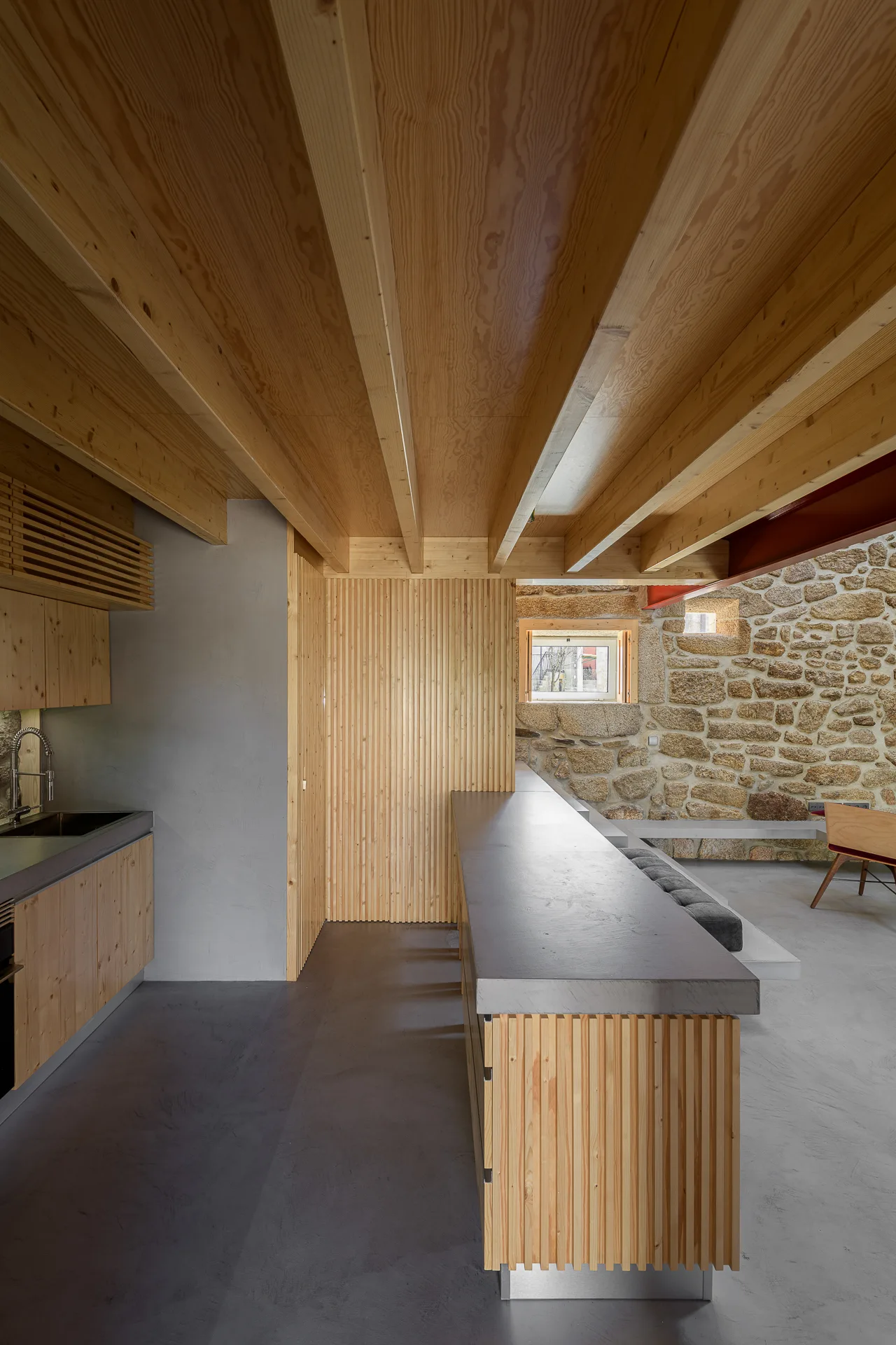 A minimalist kitchen showcases a slatted wood island, a rough stone masonry wall, smooth grey concrete flooring, and exposed timber ceiling joists.