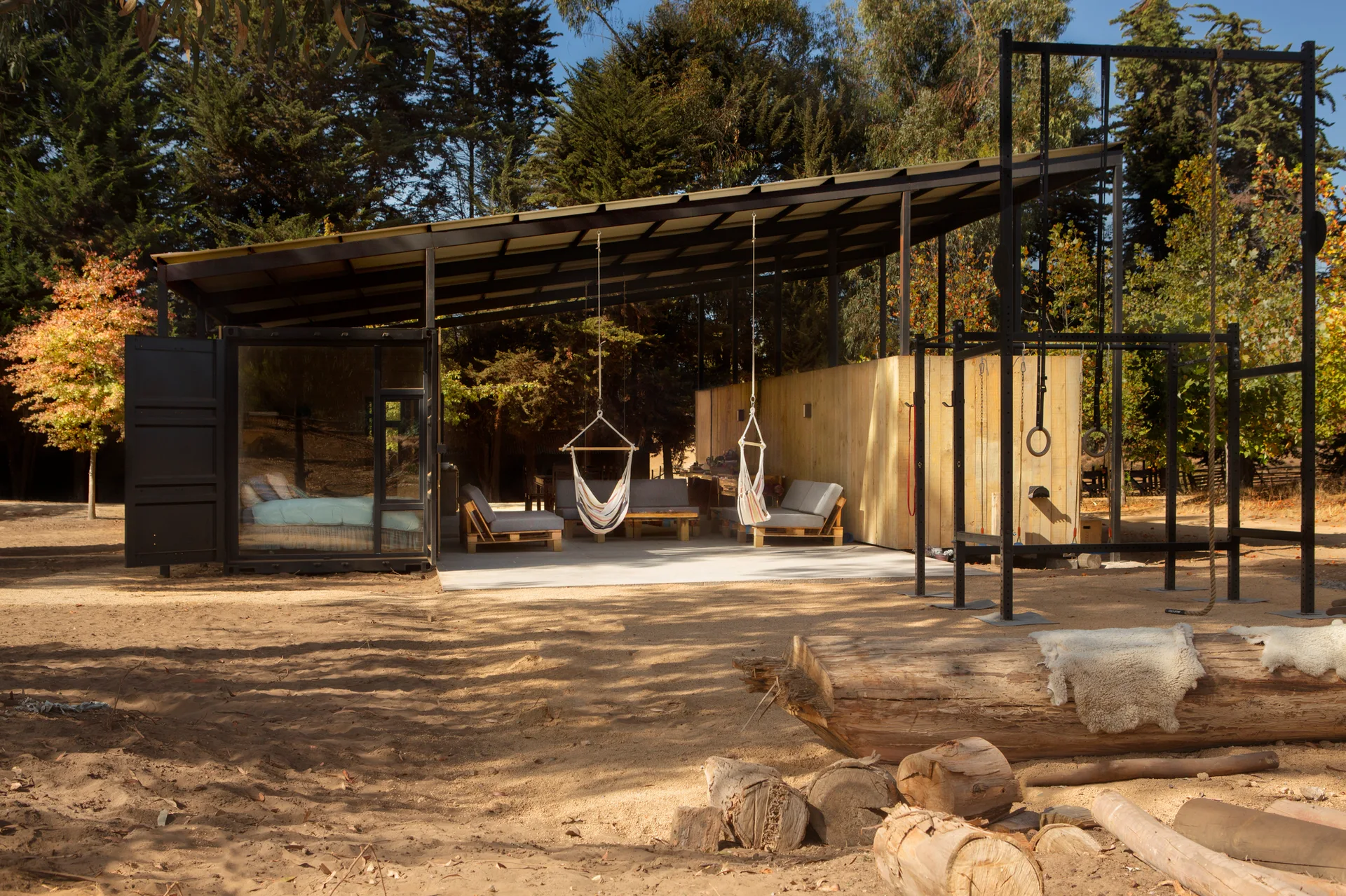 A dark shipping container cabin features an extended metal roof sheltering a concrete patio with hanging swing chairs, next to an outdoor gym rig.