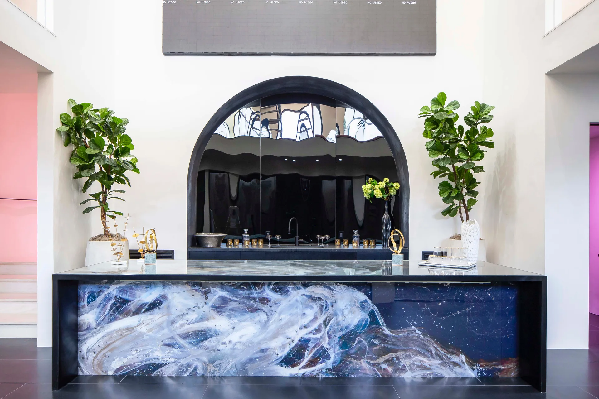 A modern bar features a glossy counter with a blue and white swirl pattern beneath a large black arched mirror flanked by two potted trees.