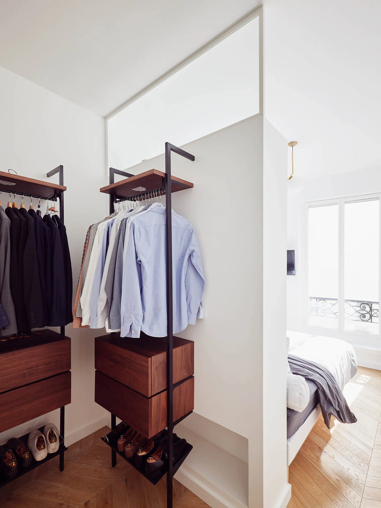 An open closet system with black metal frames and dark wood drawers stands next to a white partition wall separating it from a bright bedroom.