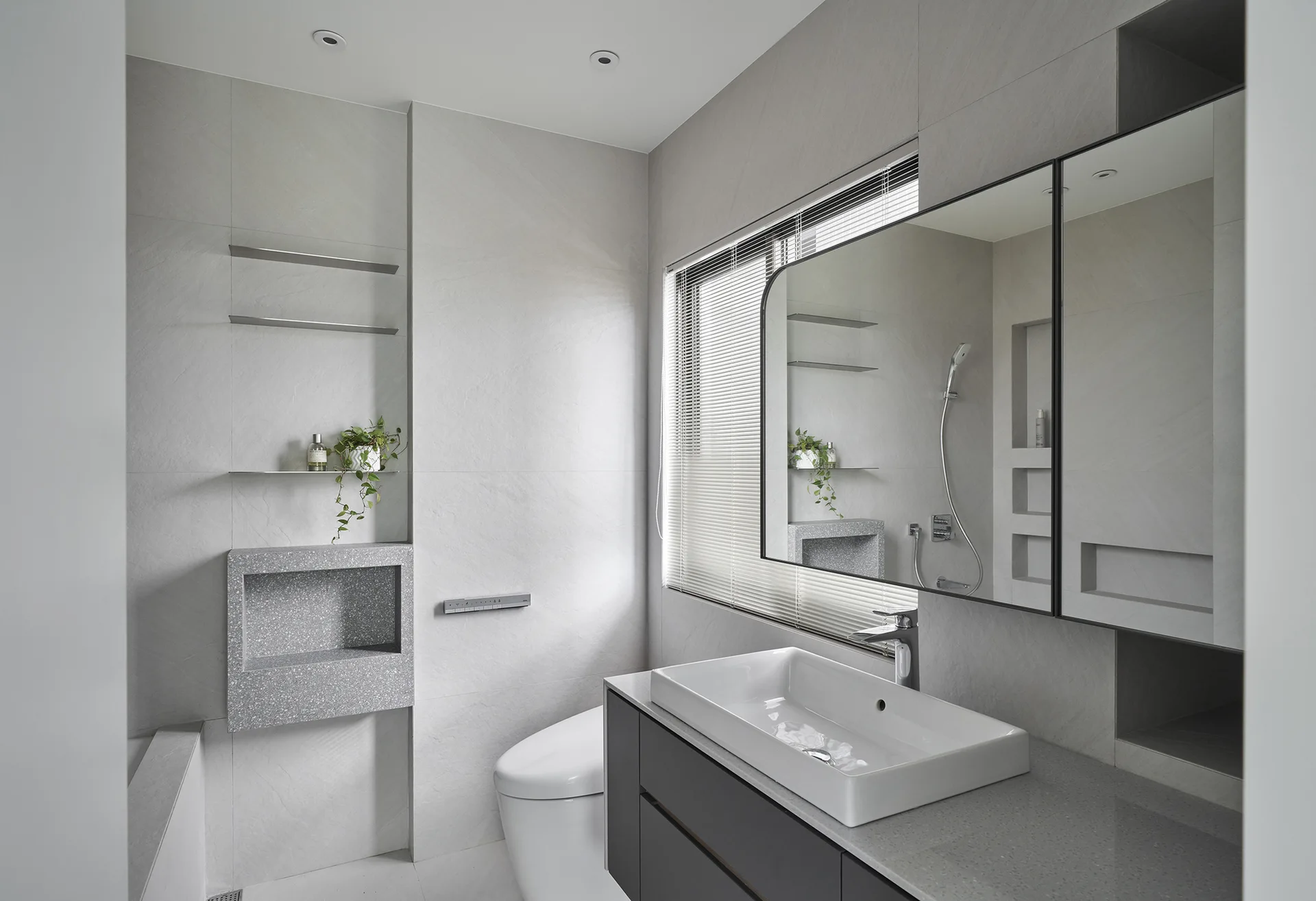 A contemporary grey bathroom featuring a dark grey vanity with a white vessel sink, a large mirror, and a speckled terrazzo wall niche.