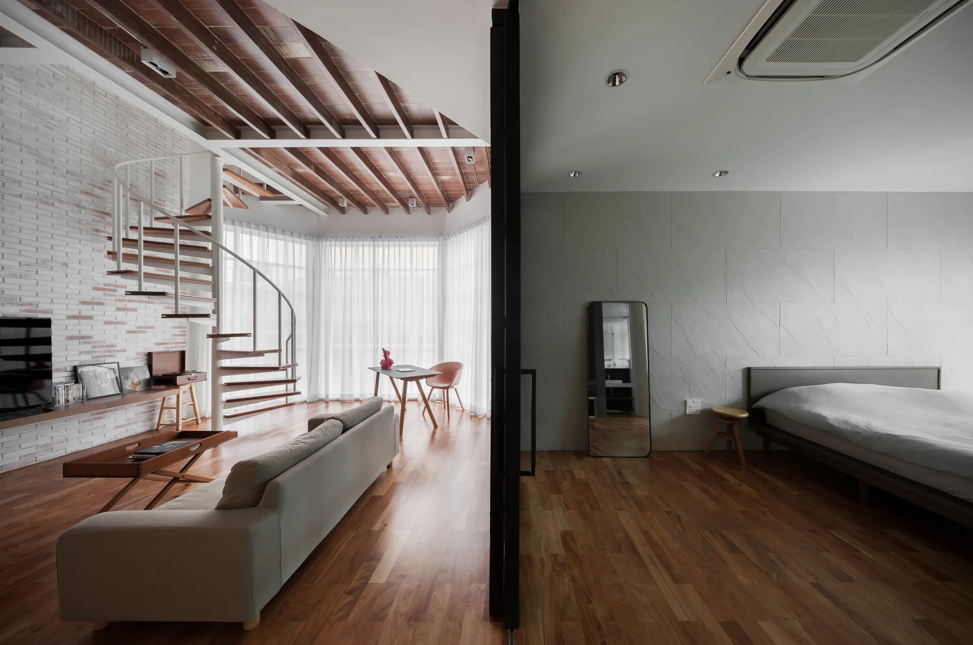 A modern loft interior split by a black-framed glass partition, showing a living room with a white spiral staircase on the left and a bedroom on the right.