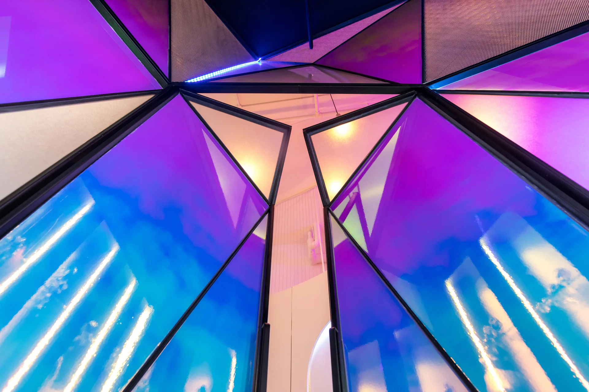 An upward view of a geometric ceiling structure with black angular frames holding iridescent purple and blue glass panels.