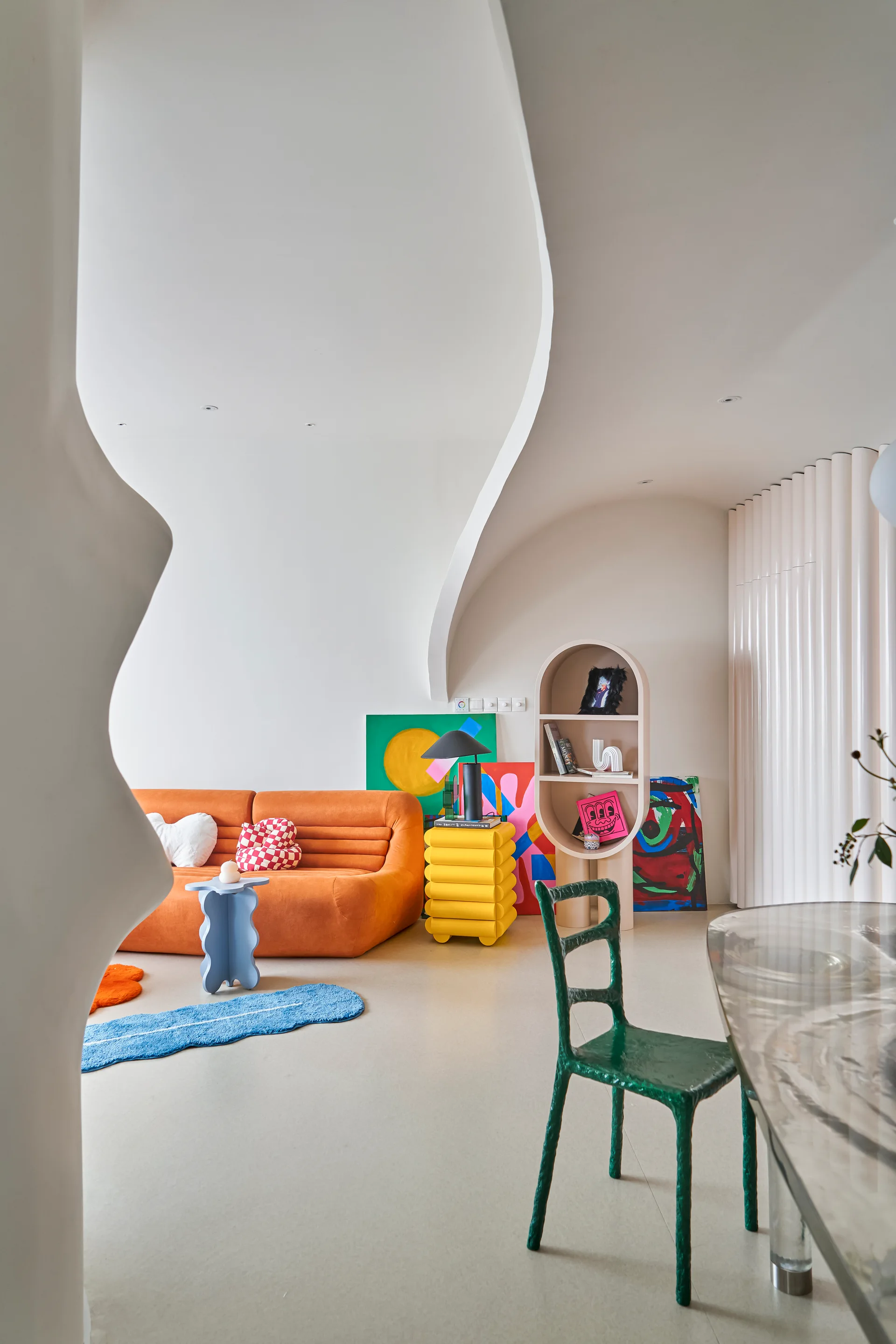 An eclectic living room features a bright orange sofa, a yellow stacked side table, a green chair, and curved white walls with abstract artwork.