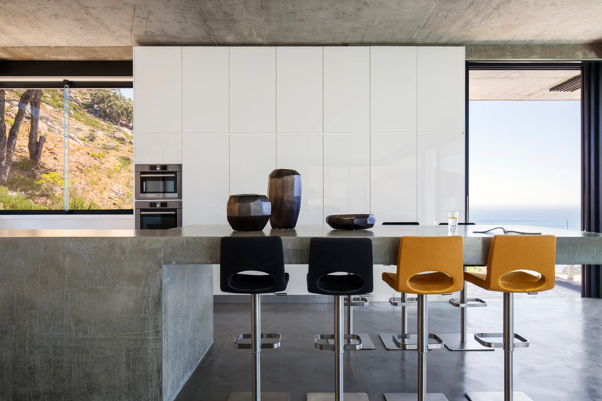 A concrete kitchen island with black and yellow stools stands before a wall of glossy white cabinets under an exposed concrete ceiling.