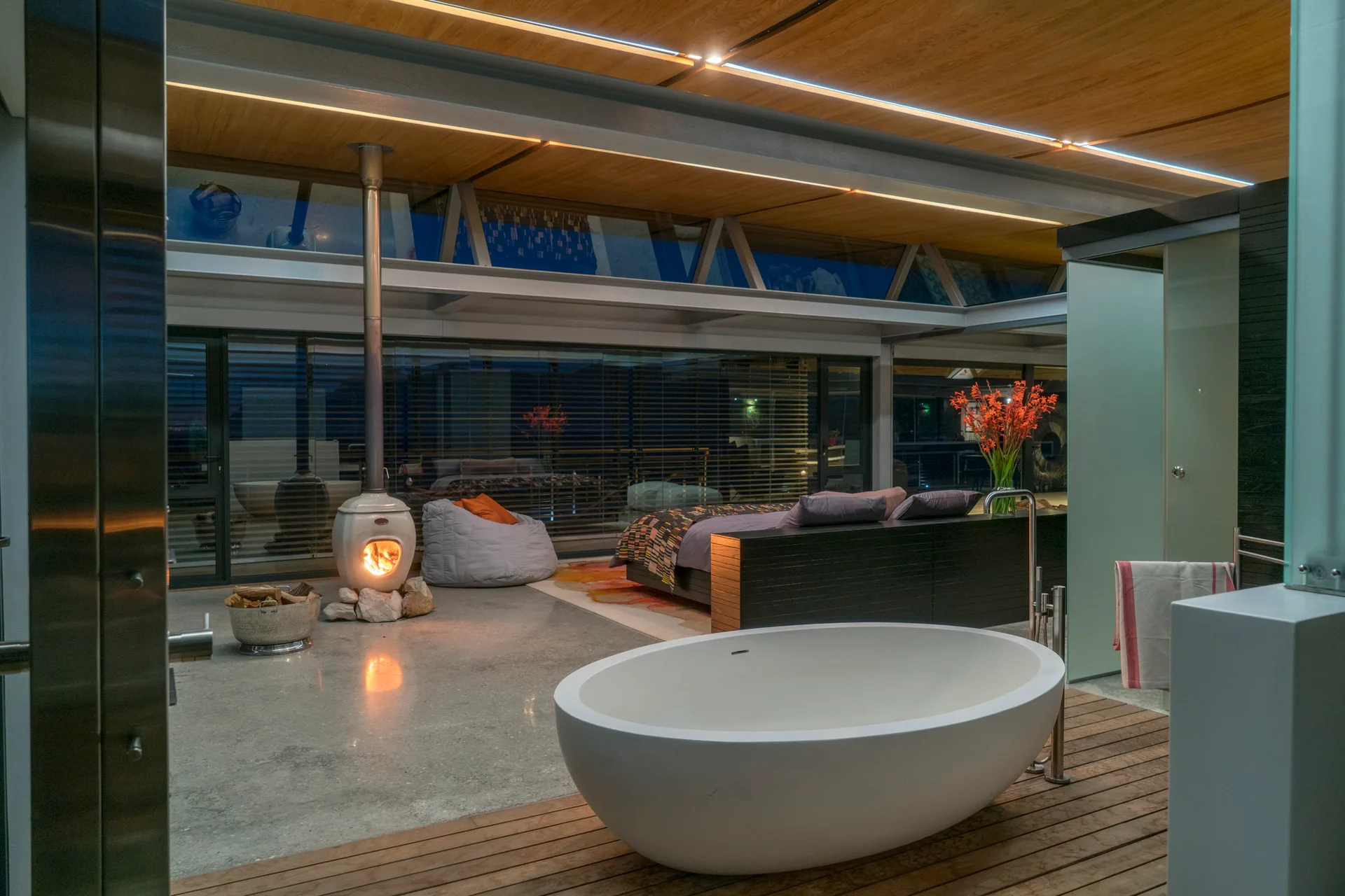 A white freestanding bathtub sits on wood decking in an open-concept bedroom containing a small modern fireplace and large glass windows.