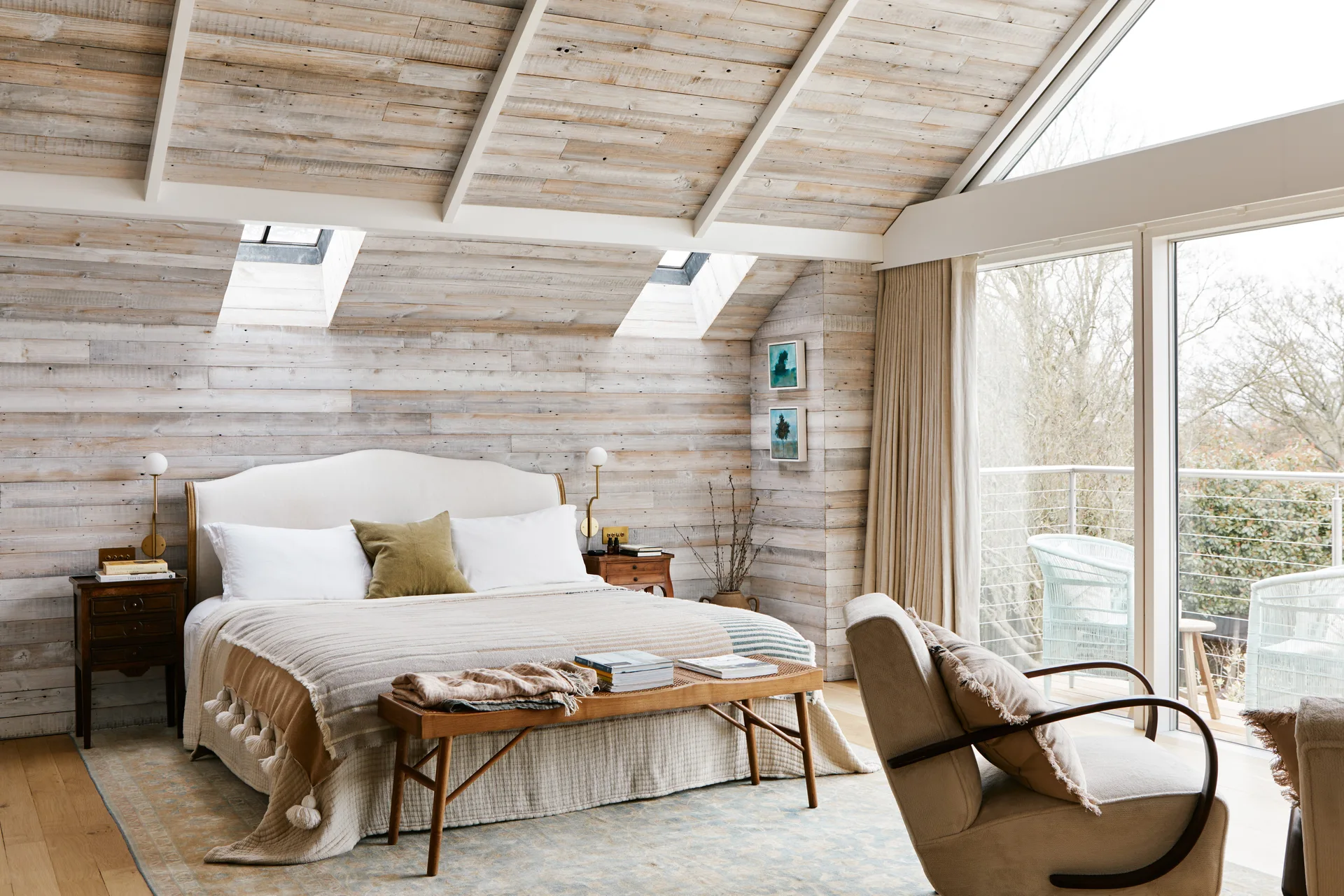 An attic bedroom features horizontal weathered wood wall paneling, a gabled ceiling with skylights, an upholstered bed, and a wooden bench.