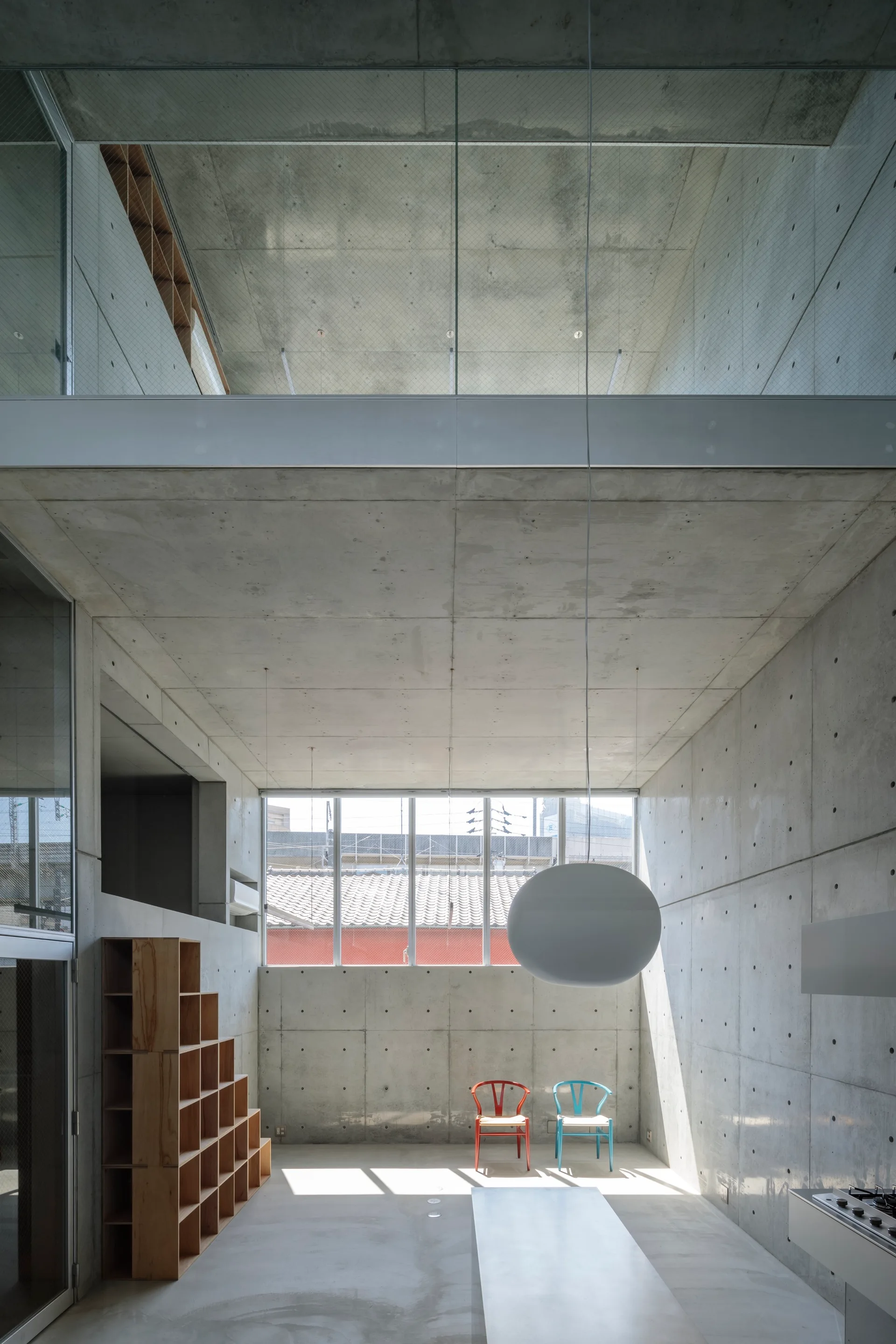 A minimalist double-height room features raw concrete walls, a mezzanine level, a large white globe pendant light, and two red and blue chairs.