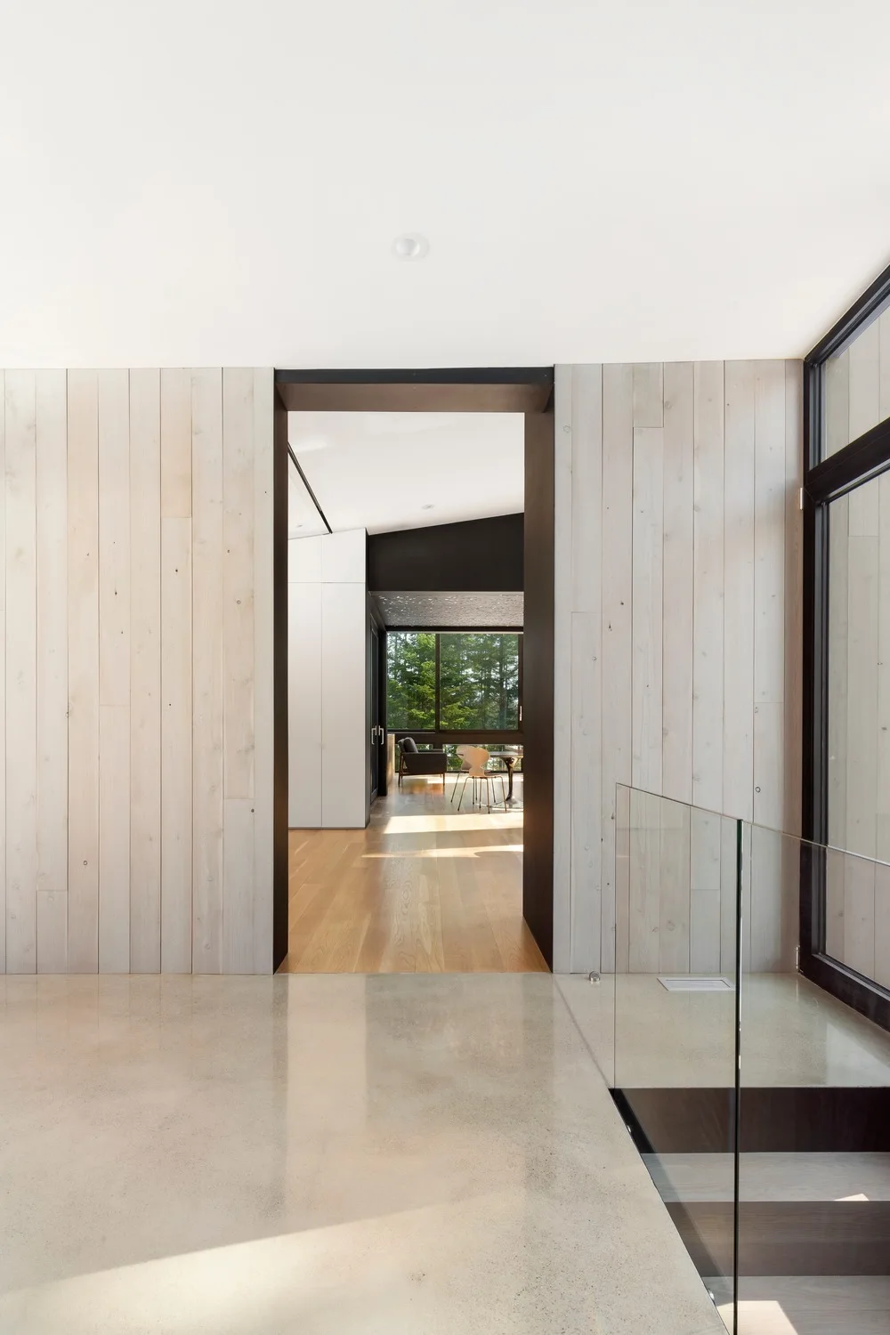 A modern hallway features vertical light wood paneled walls, a polished concrete floor, and a glass staircase railing next to a large doorway.