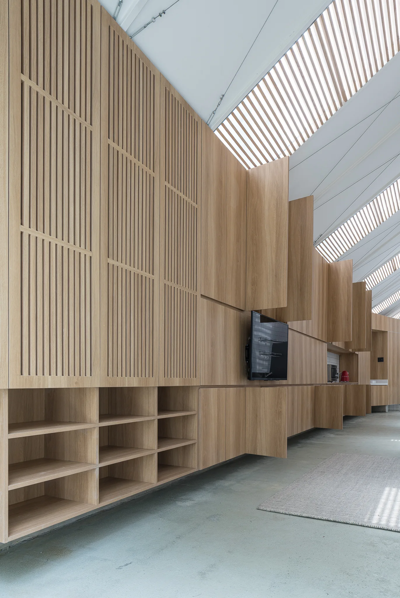 A long wall of custom light-wood cabinets featuring vertical slatted panels, open shelving, and a mounted TV under a sloped skylight ceiling.