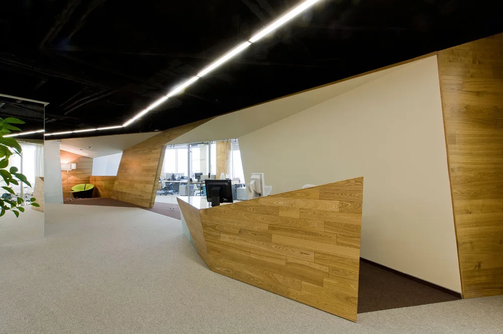 A modern office reception area featuring an angular wood desk, beige carpeted floor, and a black ceiling with a linear light strip.