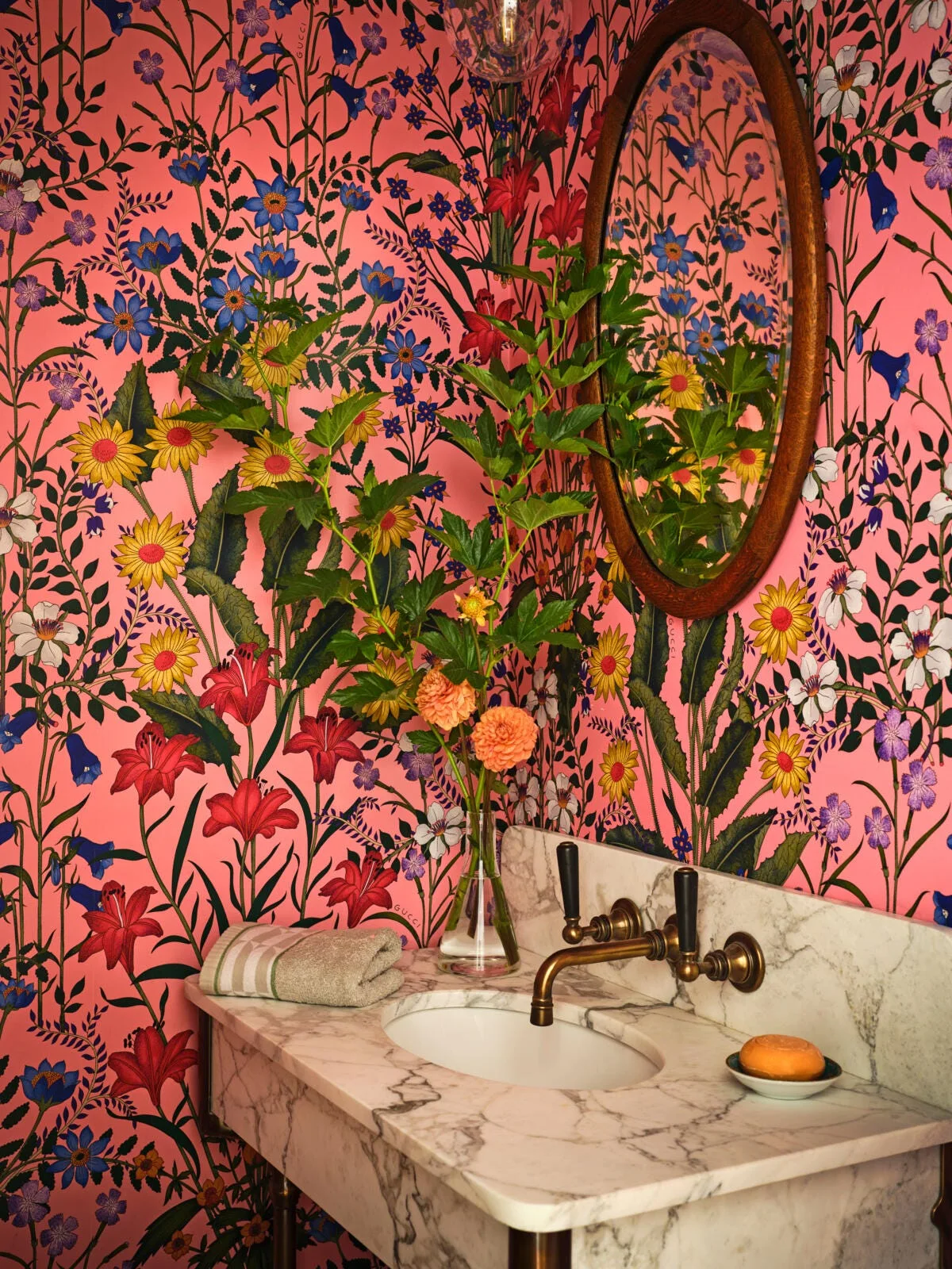 A powder room features bright pink floral wallpaper, a white marble vanity with a brass faucet, and an oval wood-framed mirror.