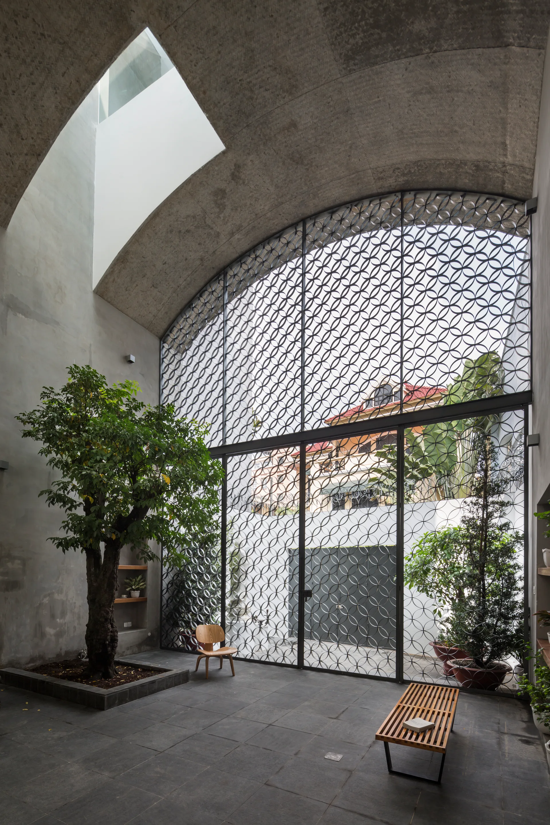 A modern indoor courtyard features a vaulted concrete ceiling with a skylight, an indoor tree, and a large arched metal screen.