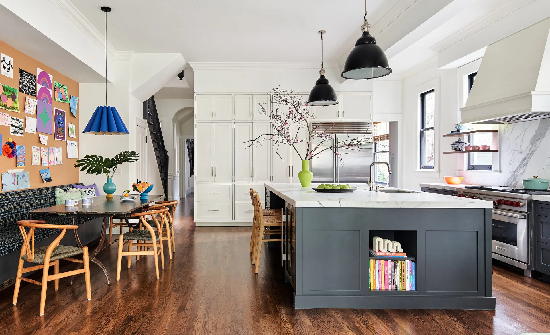 A modern kitchen features a charcoal-grey island with marble countertops, white custom cabinetry, and a dining table with built-in banquette seating.