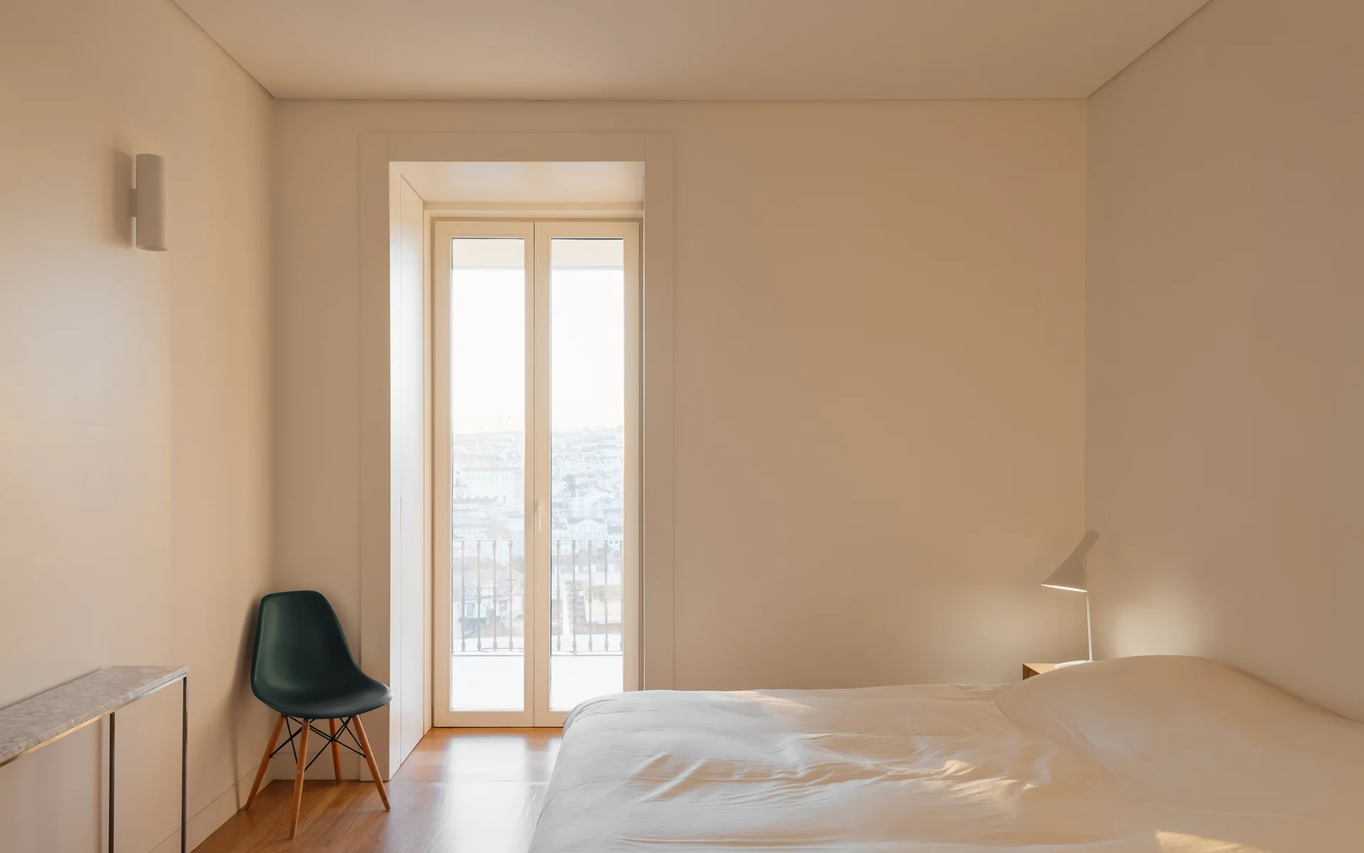 A minimalist bedroom with warm white walls features a low bed, a dark green molded plastic chair, and a tall glass door opening to a balcony.
