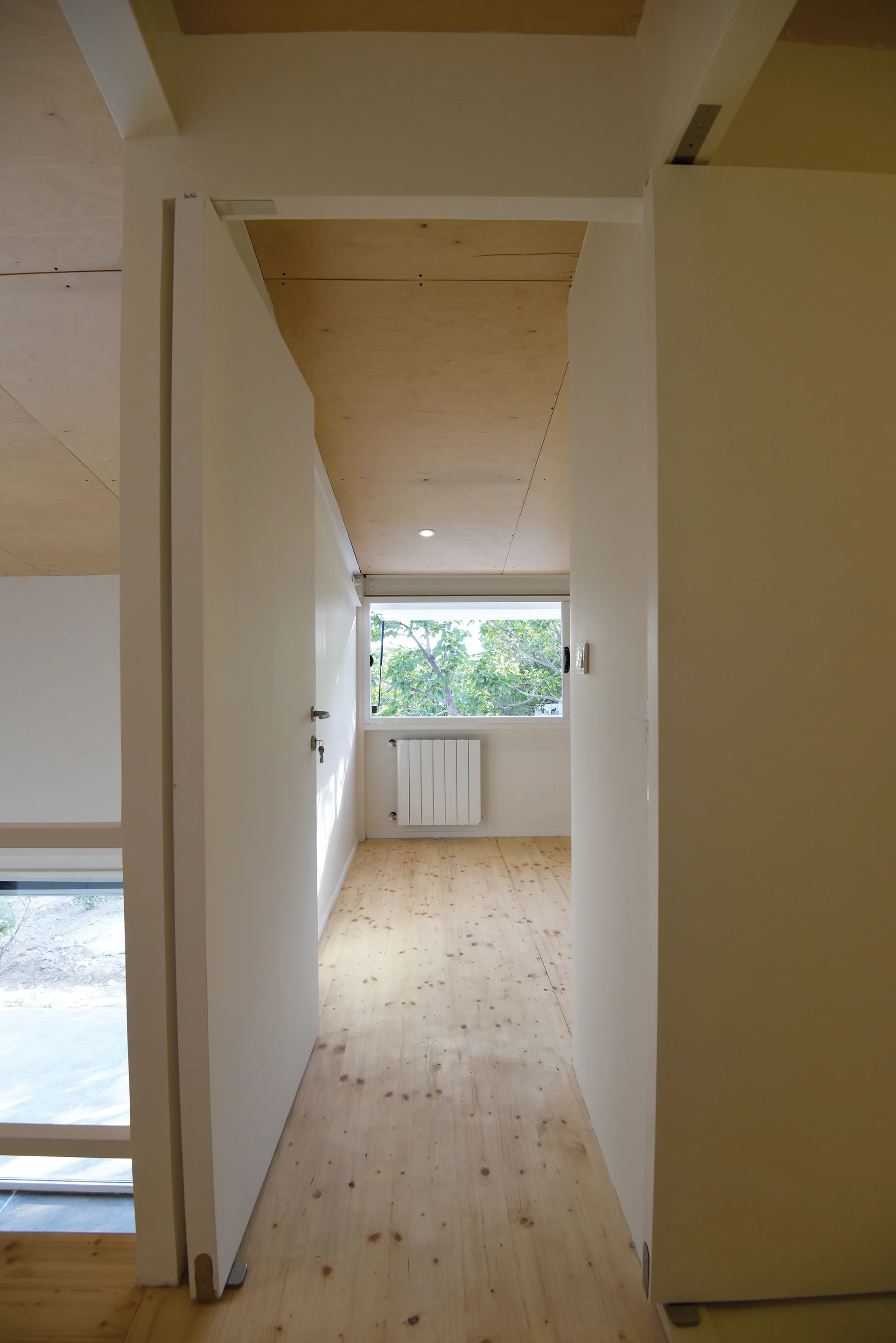 A narrow hallway features light pine wood flooring, white walls with tall flush doors, and a natural plywood ceiling leading to a window.