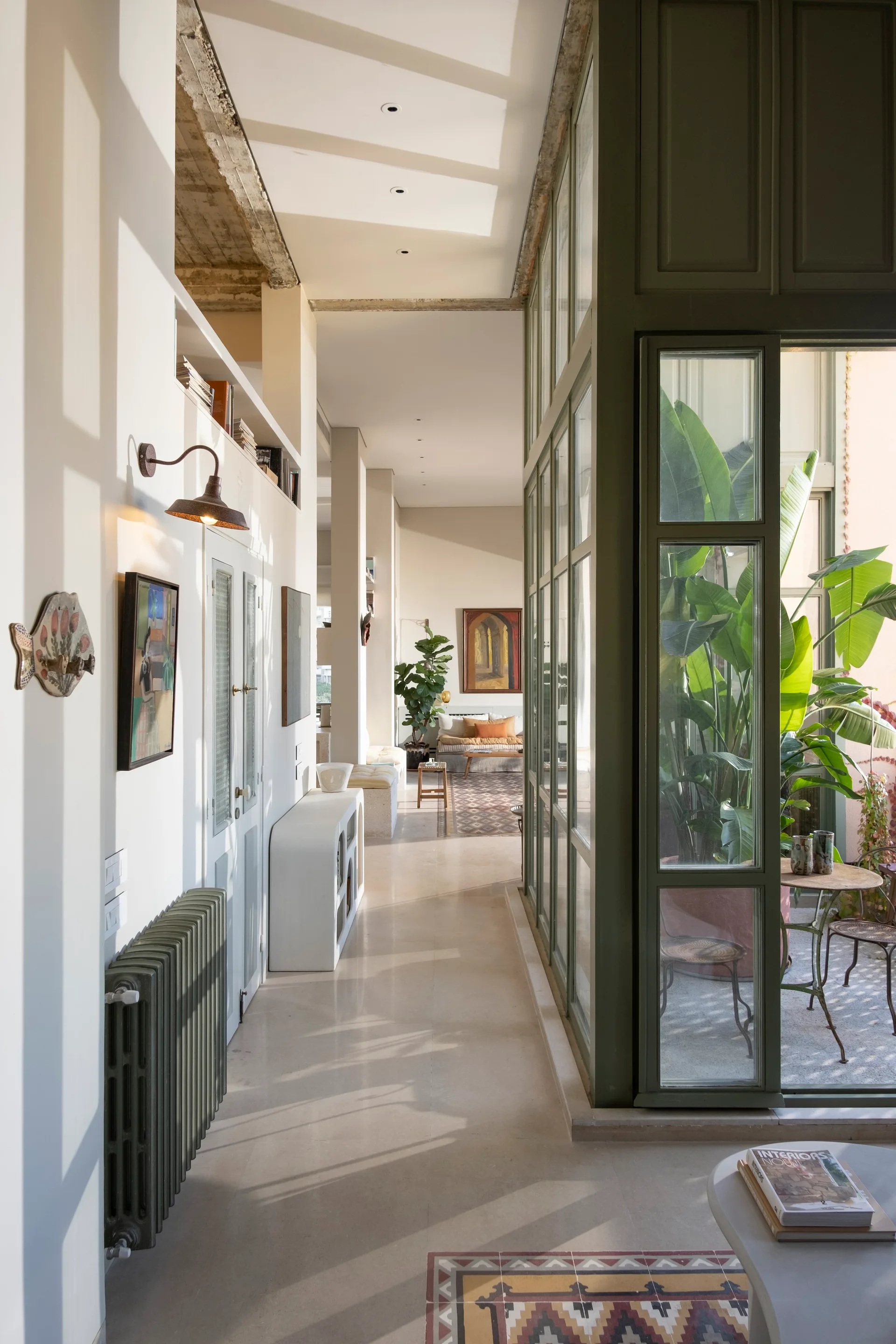 A sunlit hallway features light stone flooring, white walls, a green radiator, and a green metal-framed glass partition looking into a courtyard.