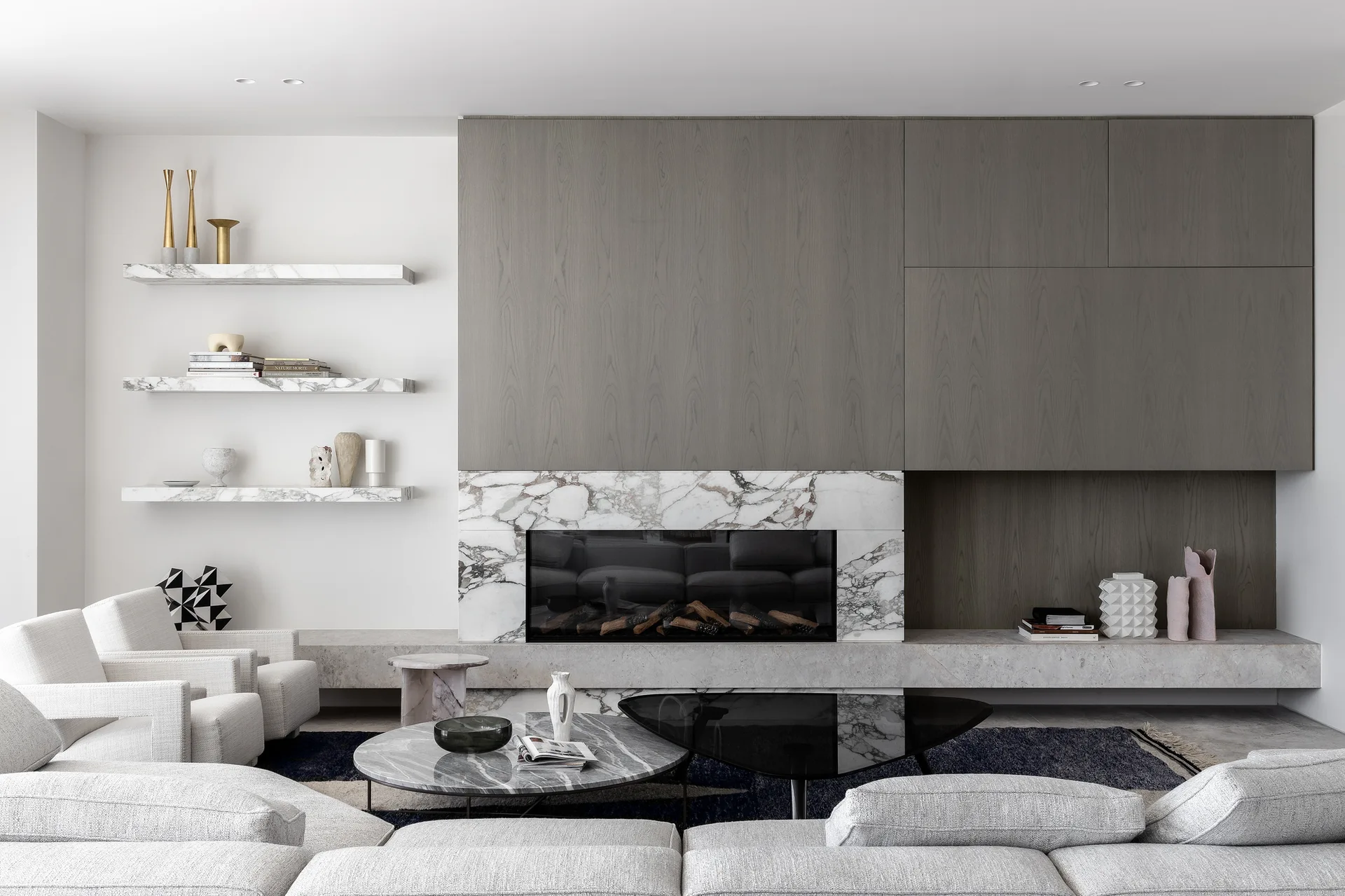 A contemporary living room features a grey wood paneled wall above a white marble fireplace, a low grey sectional sofa, and floating marble shelves.