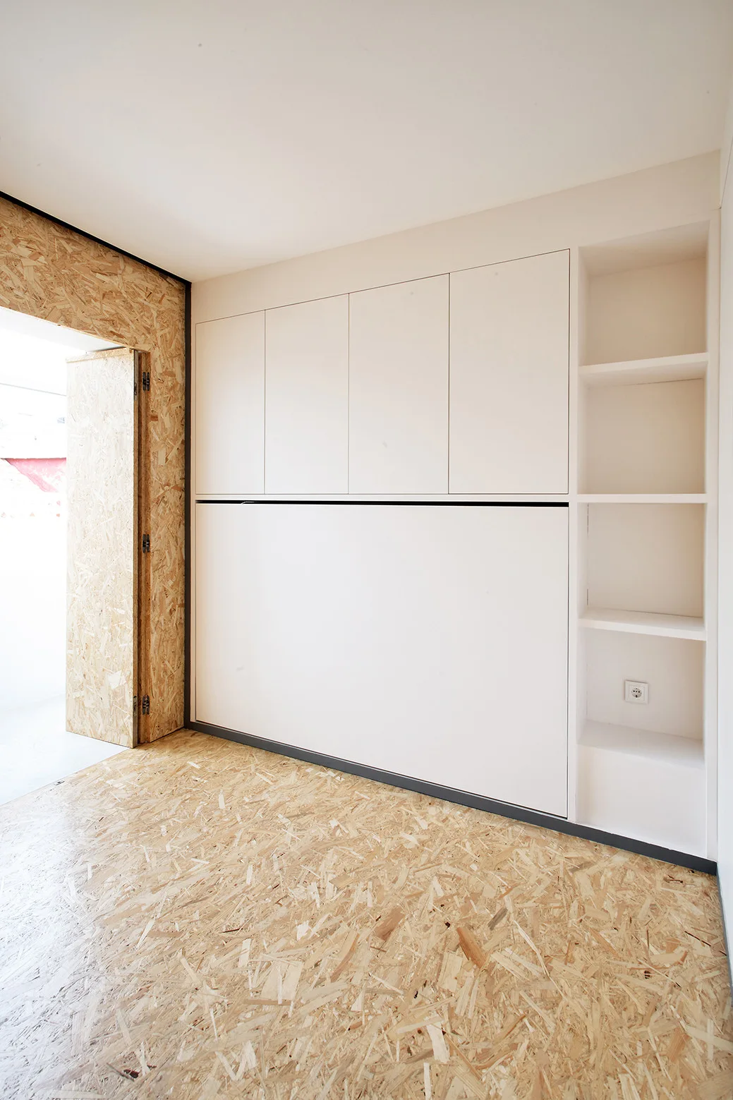A minimalist room featuring a white built-in wall cabinet with open shelving alongside an oriented strand board floor and matching door frame.
