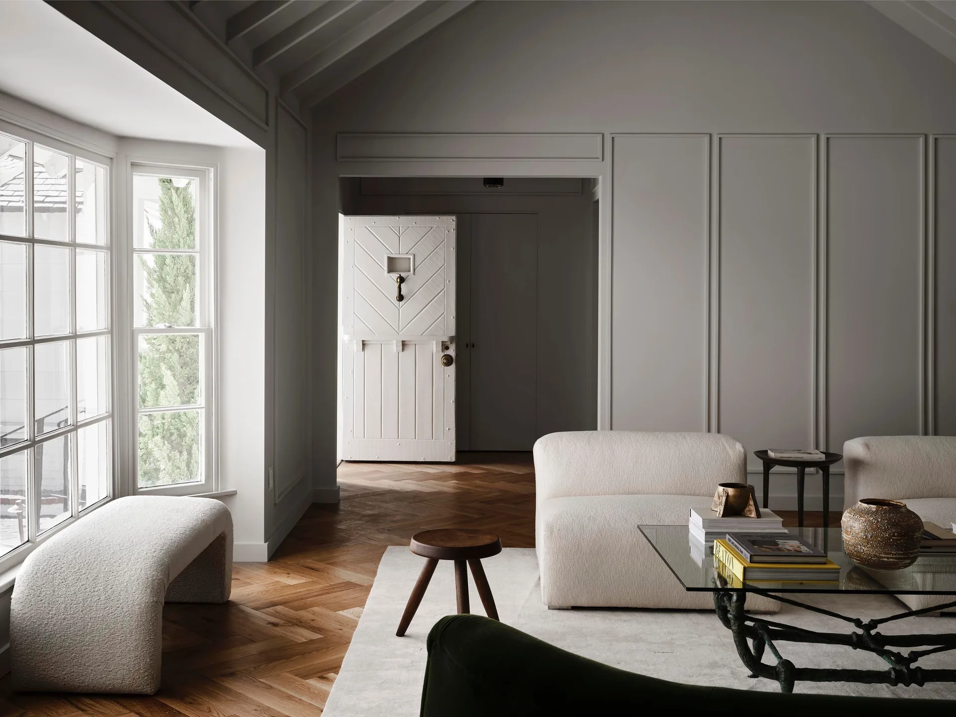 A grey living room features paneled walls, a cream upholstered sofa, a glass coffee table, and herringbone patterned wood flooring.