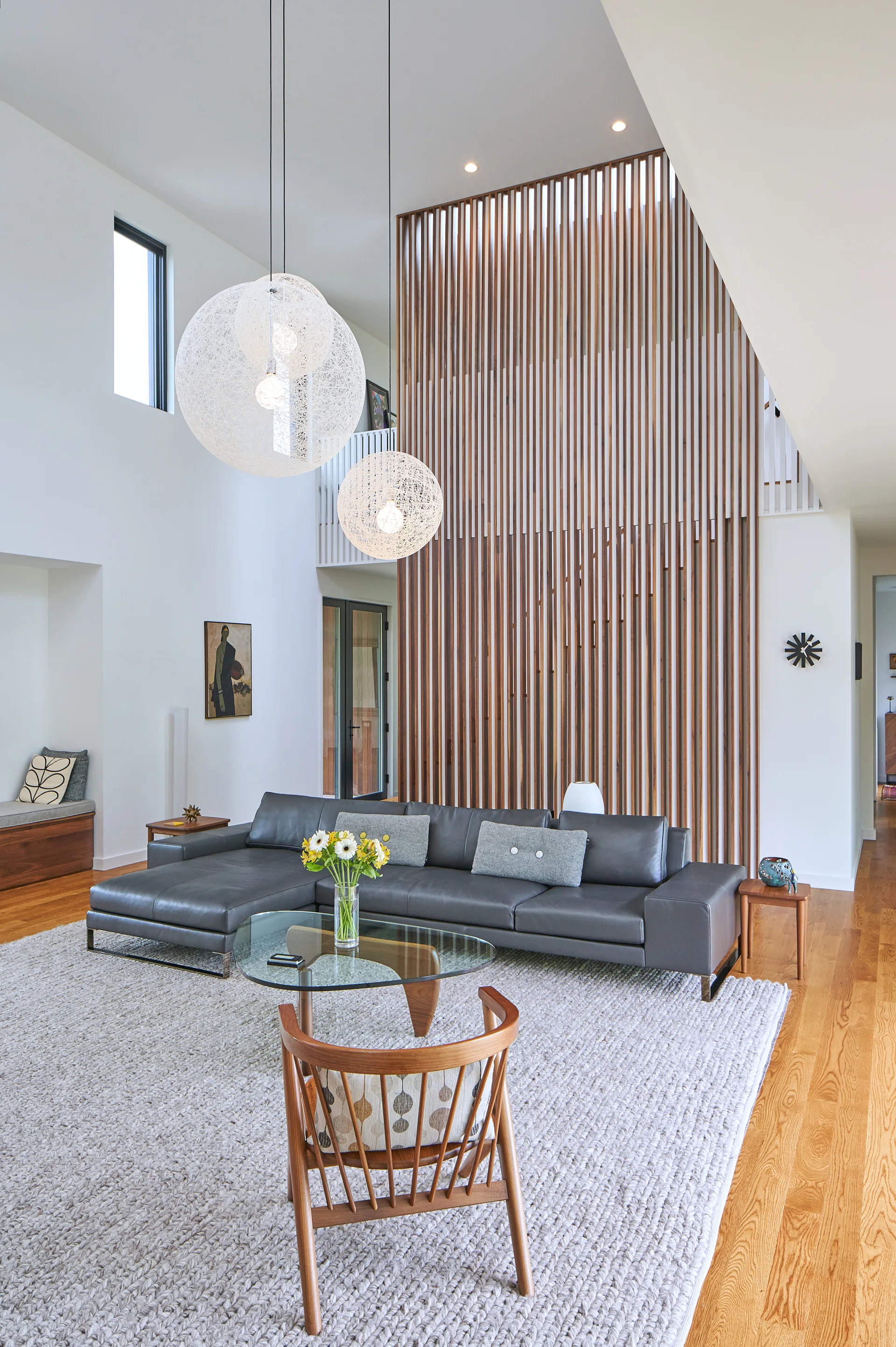A bright living room features a grey leather sectional sofa, a glass coffee table, and a tall wood slat partition wall under high ceilings.