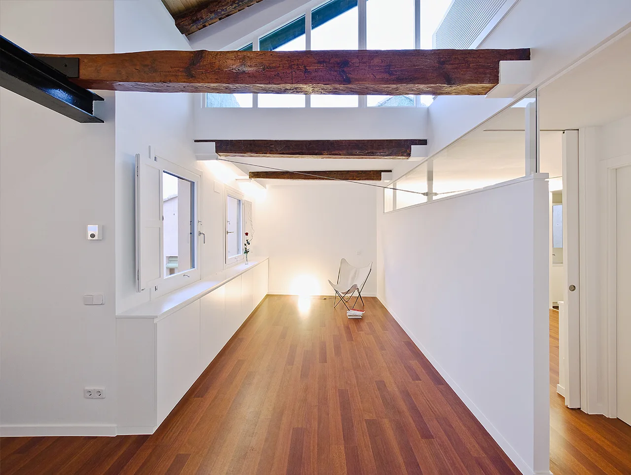 A minimalist loft hallway features warm wood floors, white walls, exposed ceiling beams, a long low cabinet, and a single white butterfly chair.