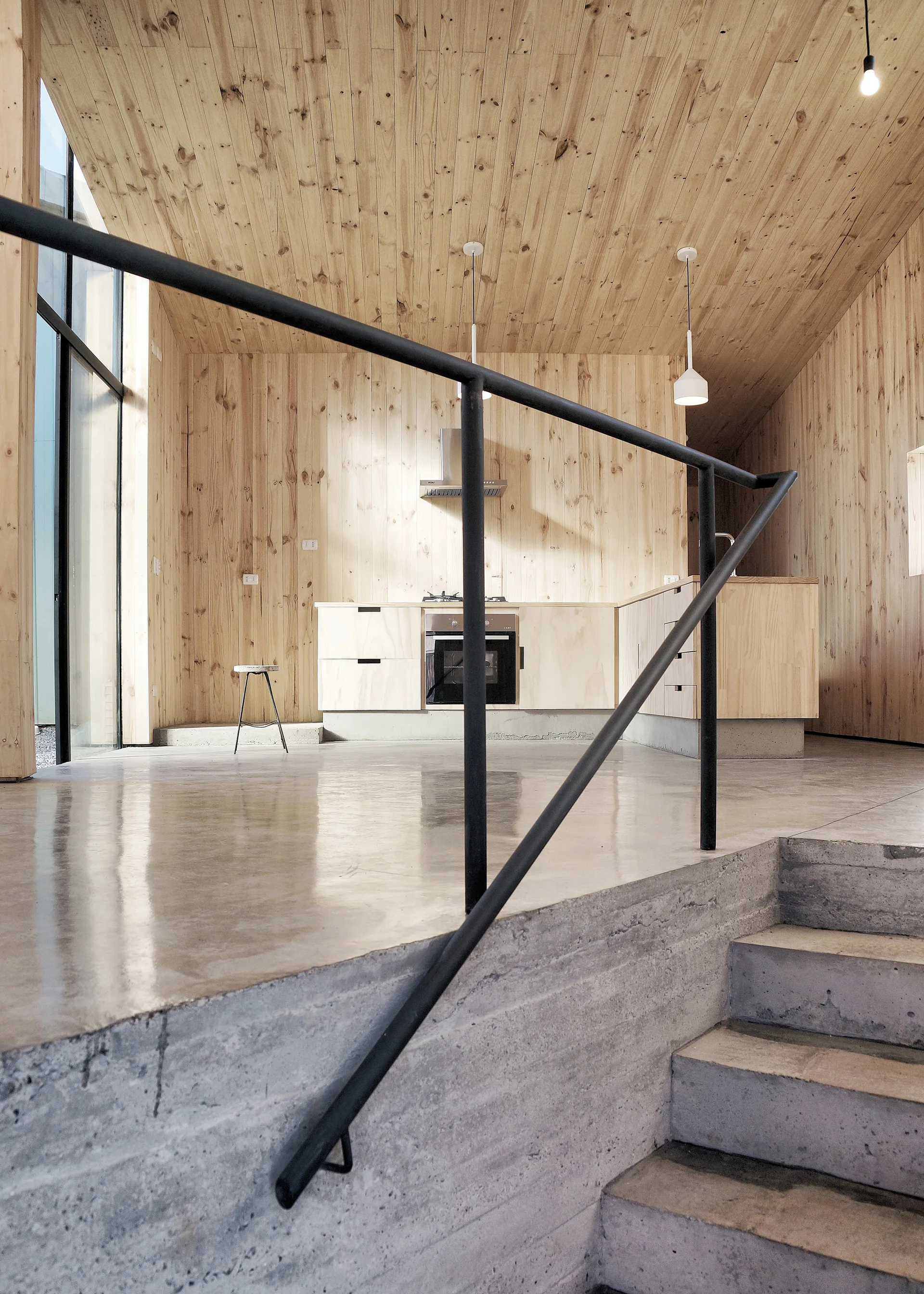 Concrete stairs with a black metal handrail lead to a split-level kitchen featuring light wood-paneled walls, matching cabinets, and polished floors.