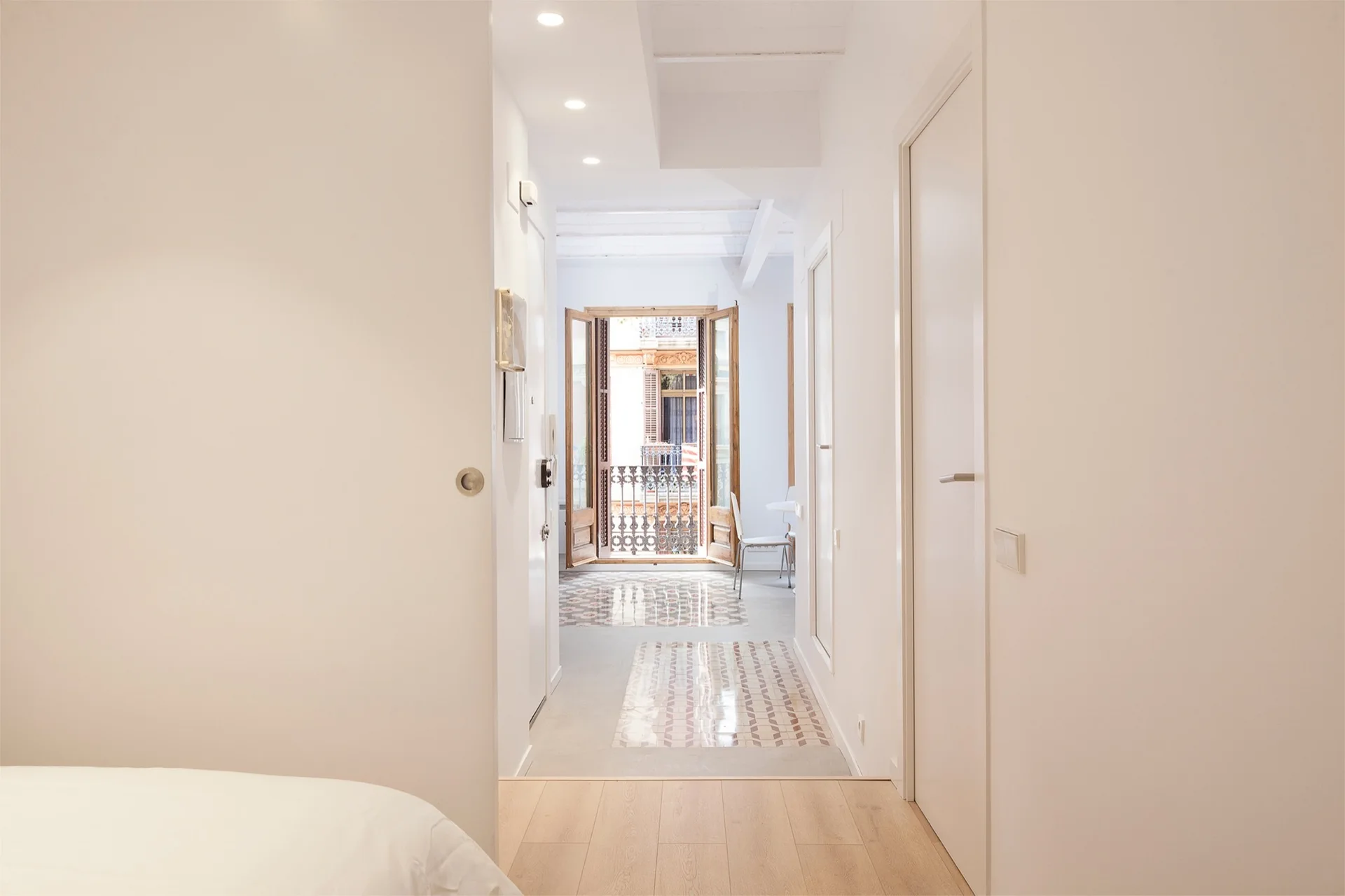 A bright white hallway featuring a transition from light wood flooring to patterned geometric floor tiles leading to an open wooden balcony door.