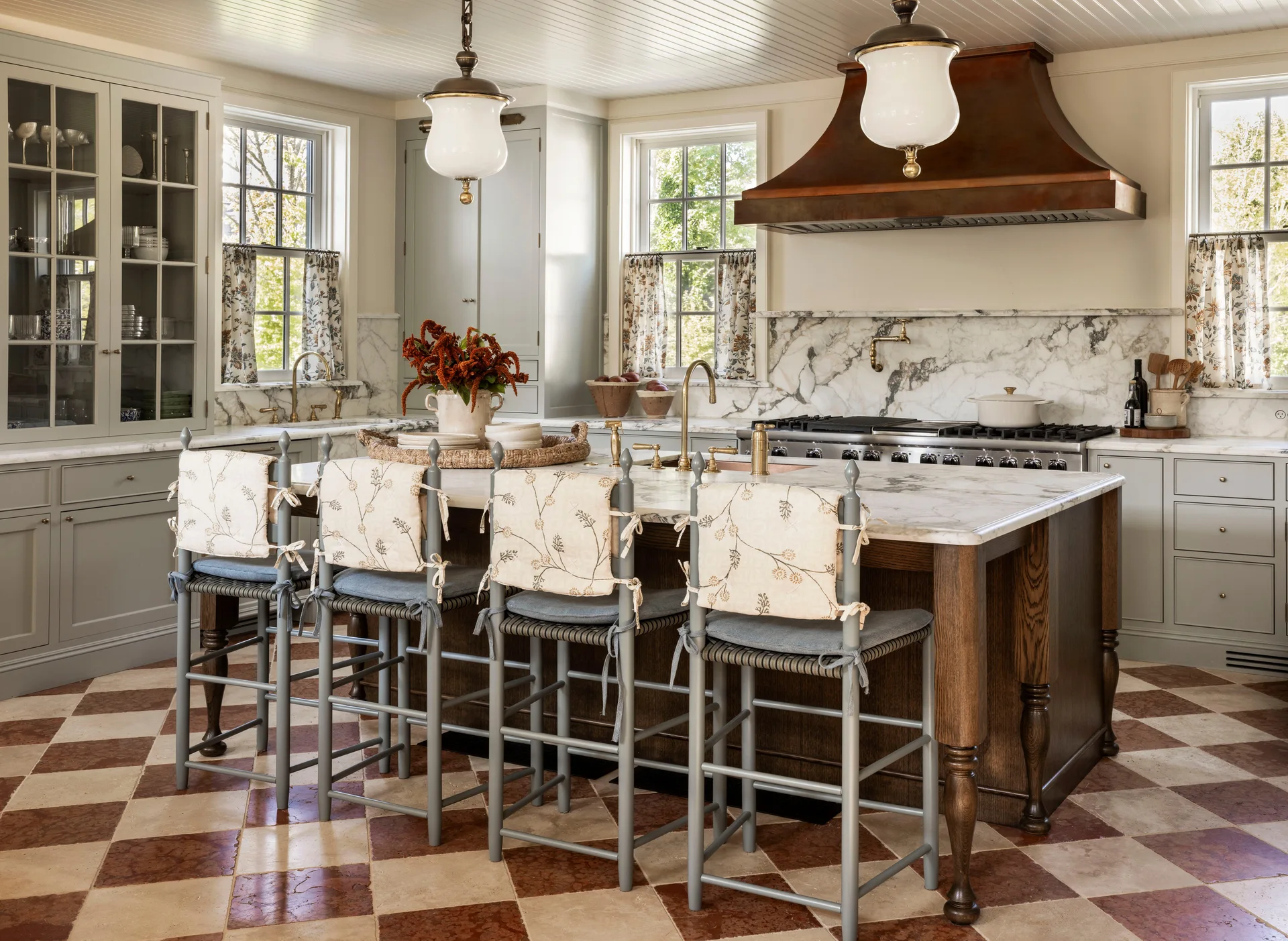A traditional kitchen features a marble-topped island with gray bar stools, a prominent copper range hood, and a checkerboard tile floor.
