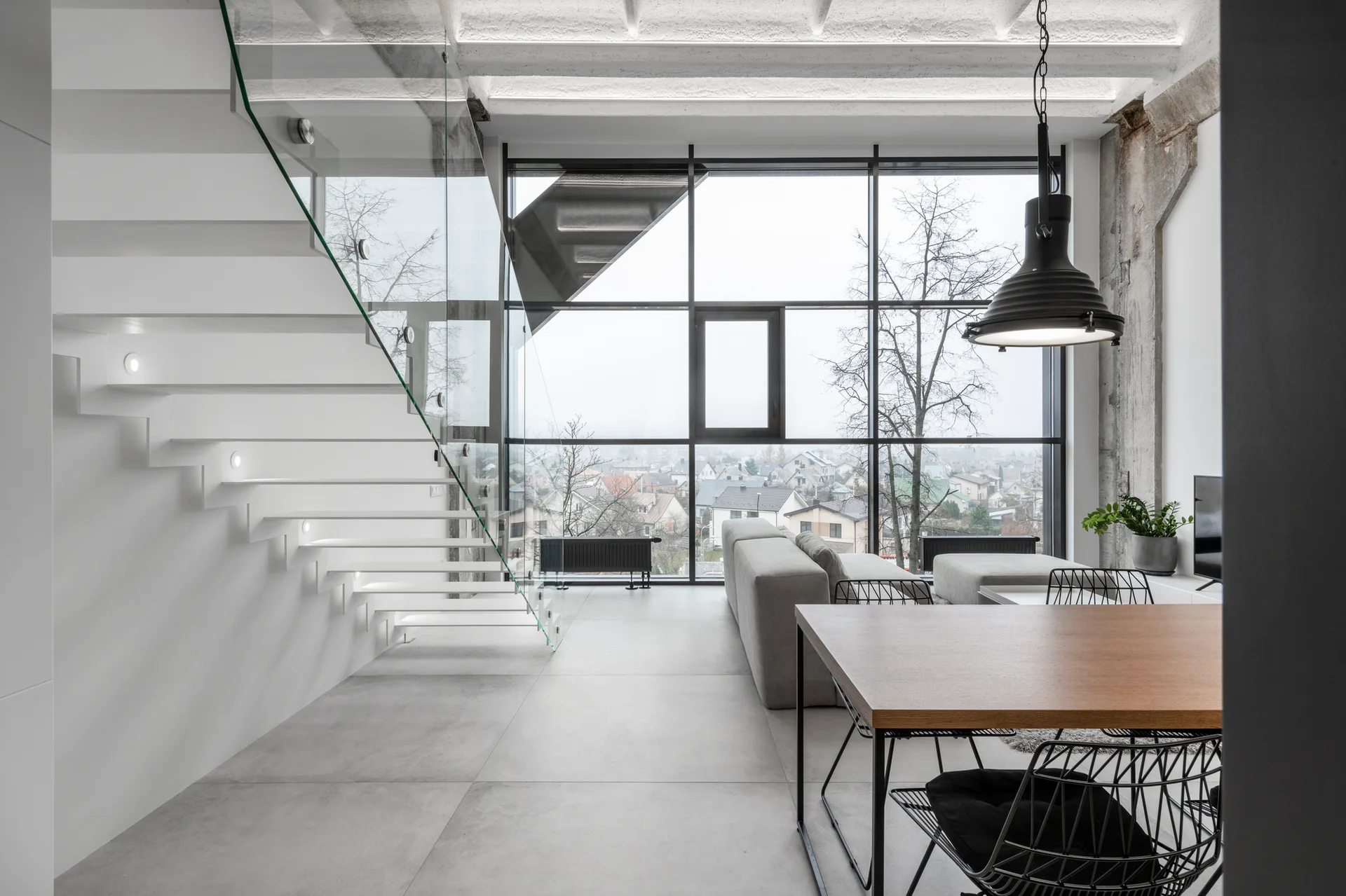 A modern living area features a white floating staircase with glass railing, a wooden dining table, and a large grid window overlooking a town.