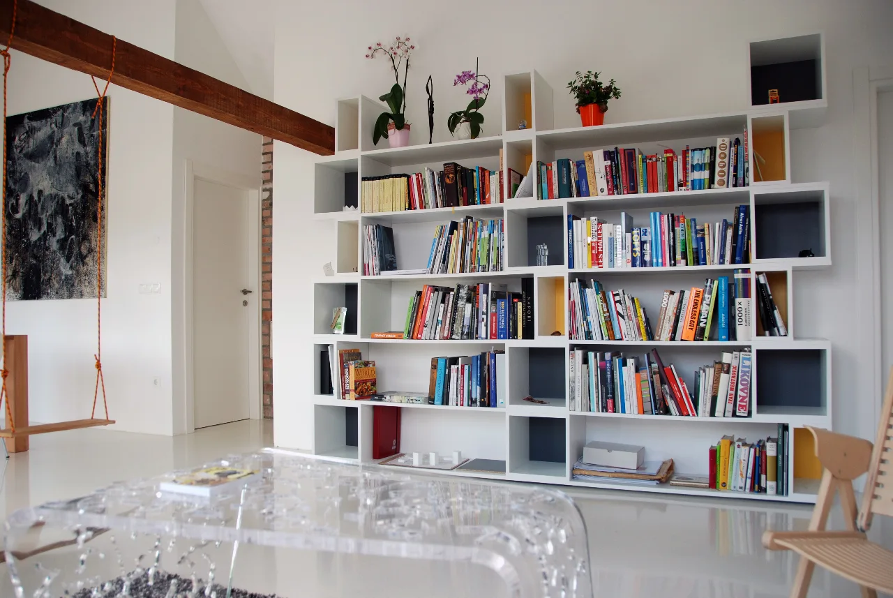 A bright living room featuring a large geometric white bookshelf filled with books, a glossy white floor, and a transparent acrylic coffee table.