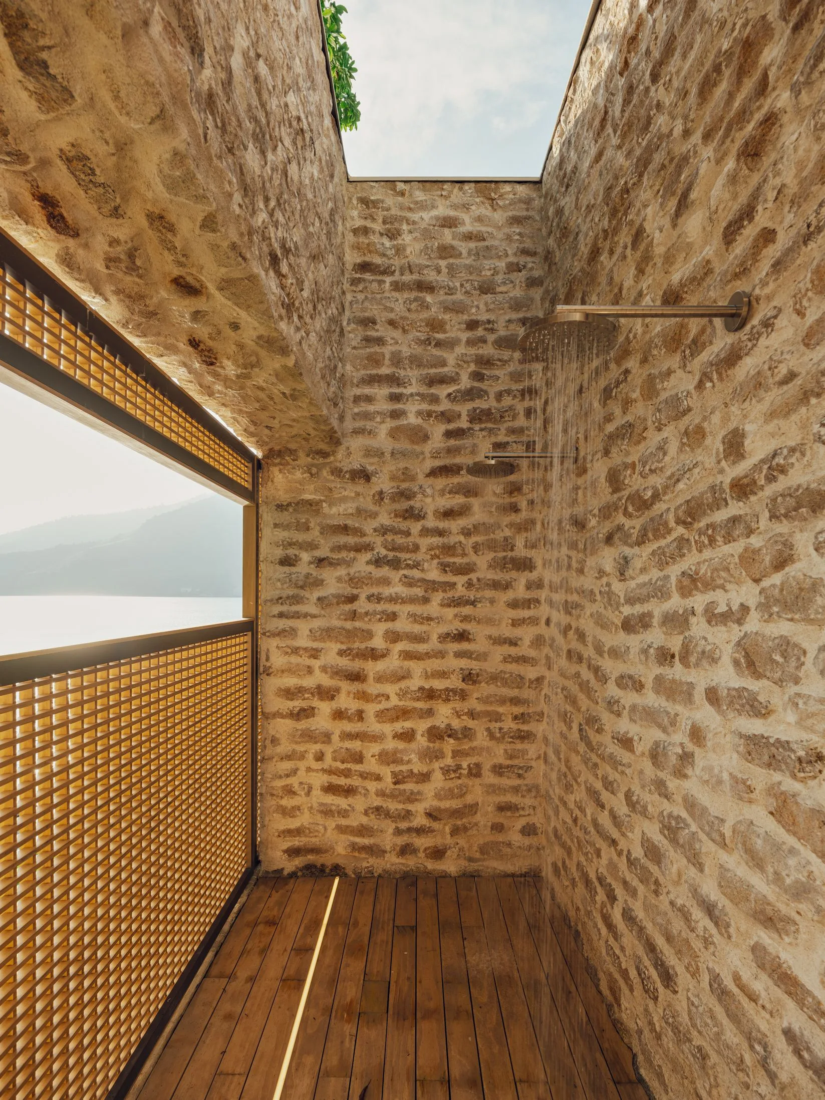 An open-air stone shower with a slatted wooden floor, a geometric lattice screen, and an open ceiling showing the sky.