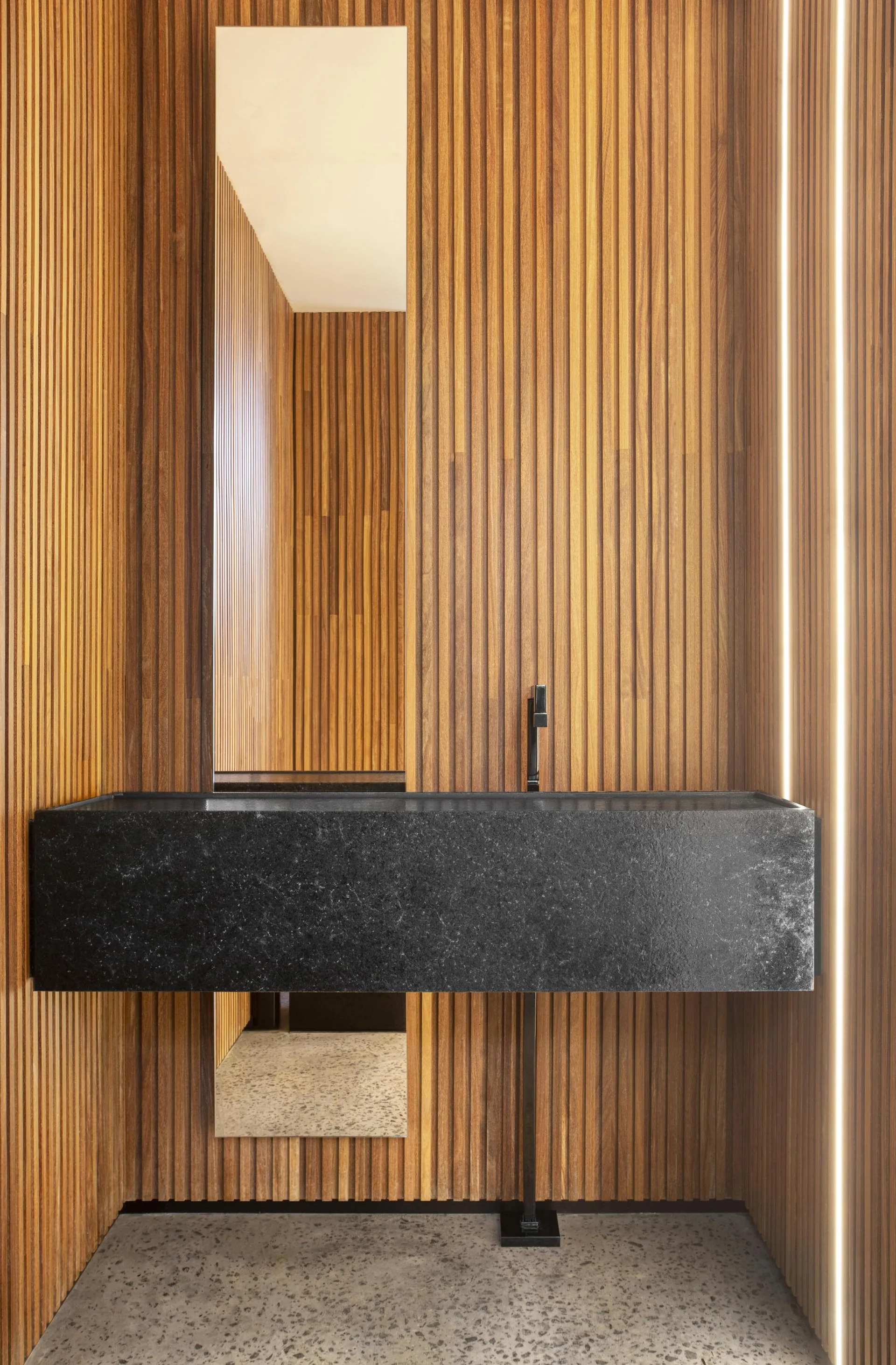 A minimalist powder room with a floating black stone basin against vertical wood slat walls featuring integrated vertical LED strip lighting.