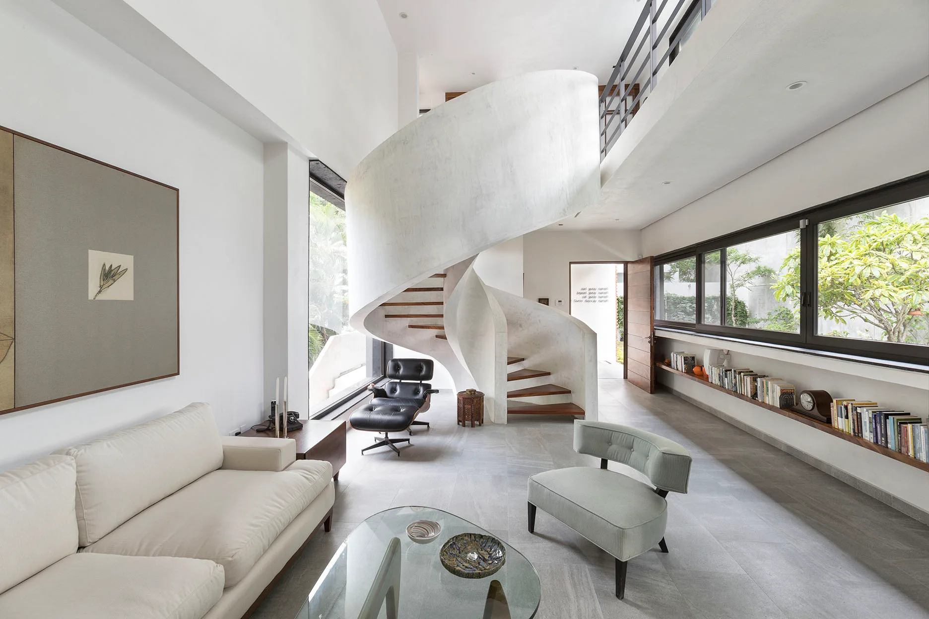 A modern living room features an off-white sofa, a sculptural white spiral staircase, and a long wooden floating bookshelf beneath a wide window.