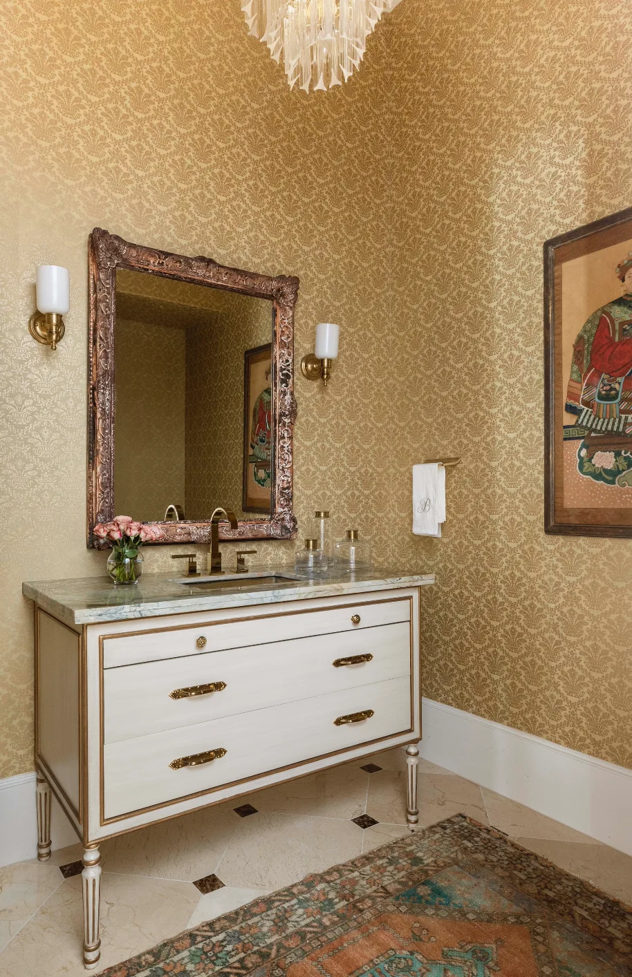 A powder room features gold damask wallpaper, a white chest-style vanity with brass hardware, an ornate gold mirror, and wall sconces.