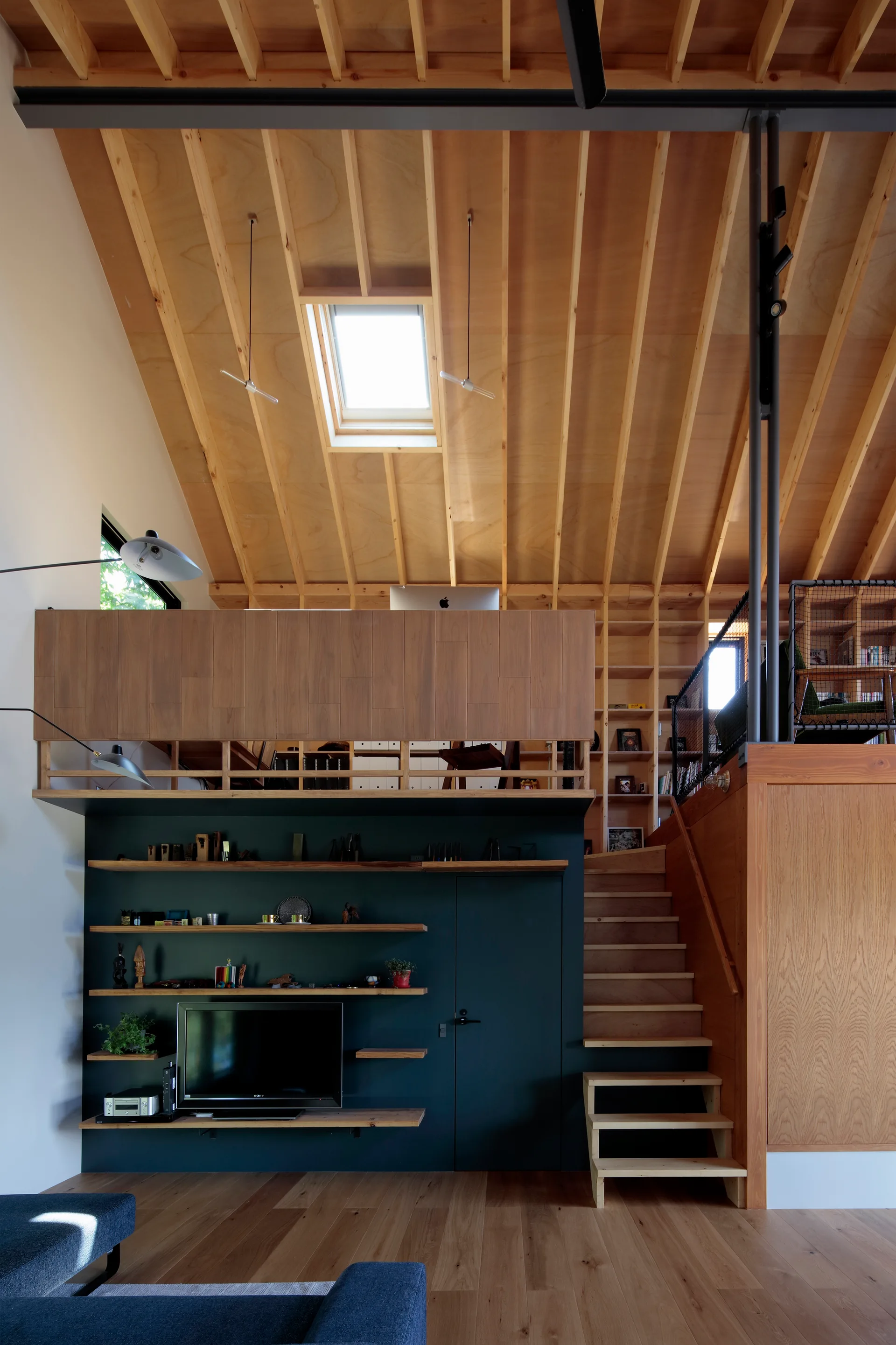 A modern living room featuring a wooden staircase leading to a mezzanine level under an exposed timber ceiling with a skylight and a dark gray accent wall.