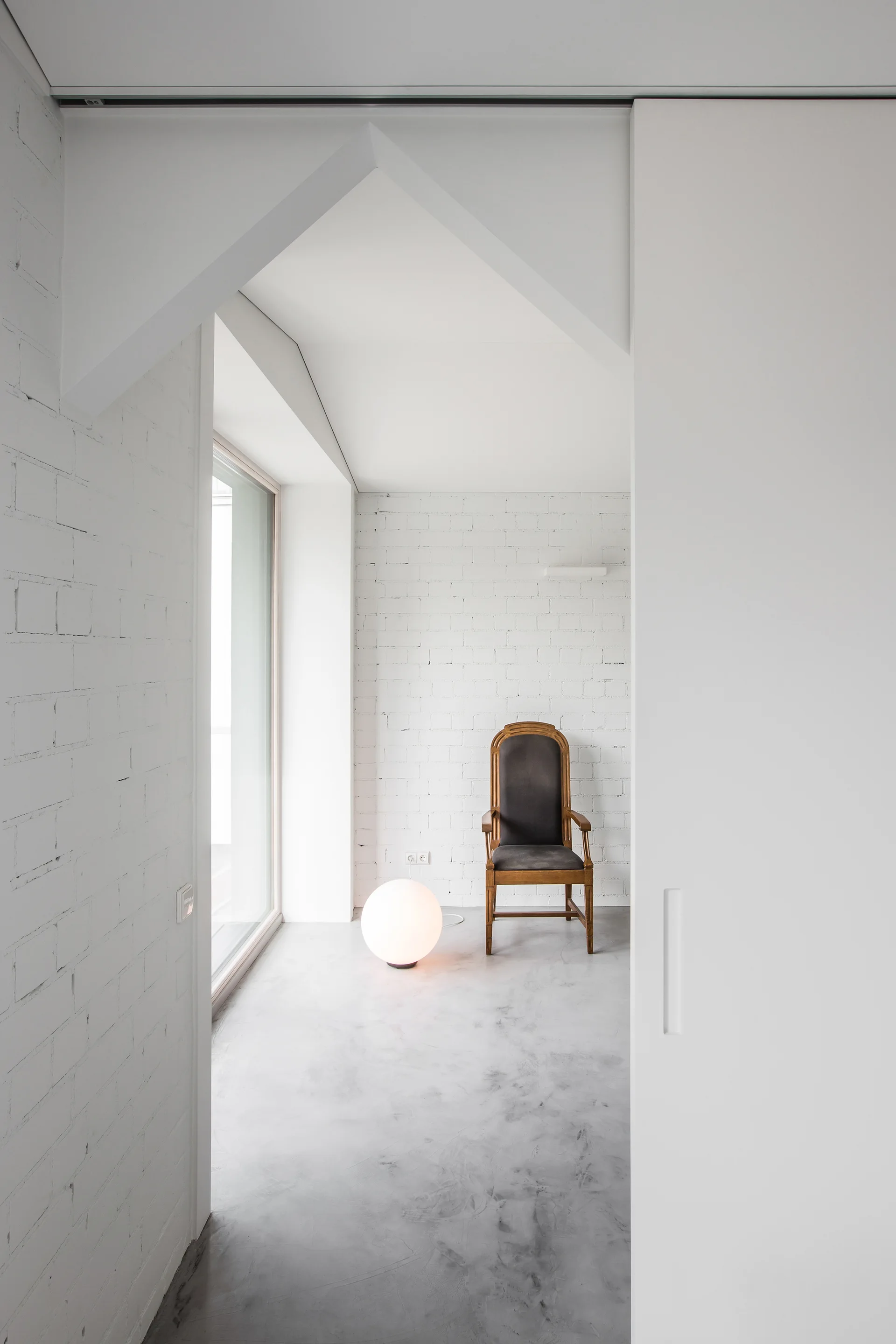 A minimalist room with white brick walls, a light gray concrete floor, a vintage wooden armchair, and a glowing globe floor lamp.