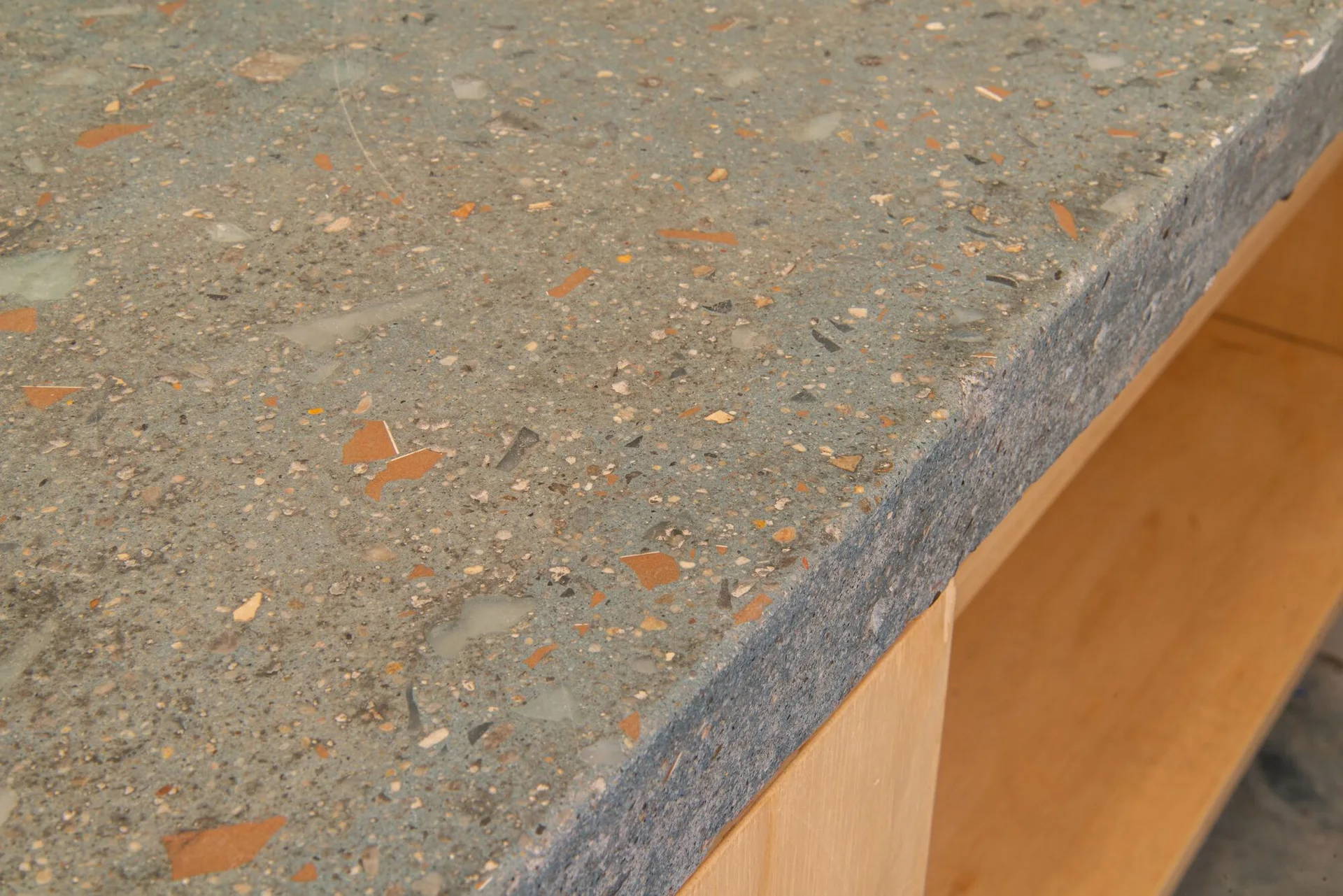 A close-up of a grey speckled terrazzo countertop with warm-toned orange and tan aggregate chips resting on a light wood cabinet.